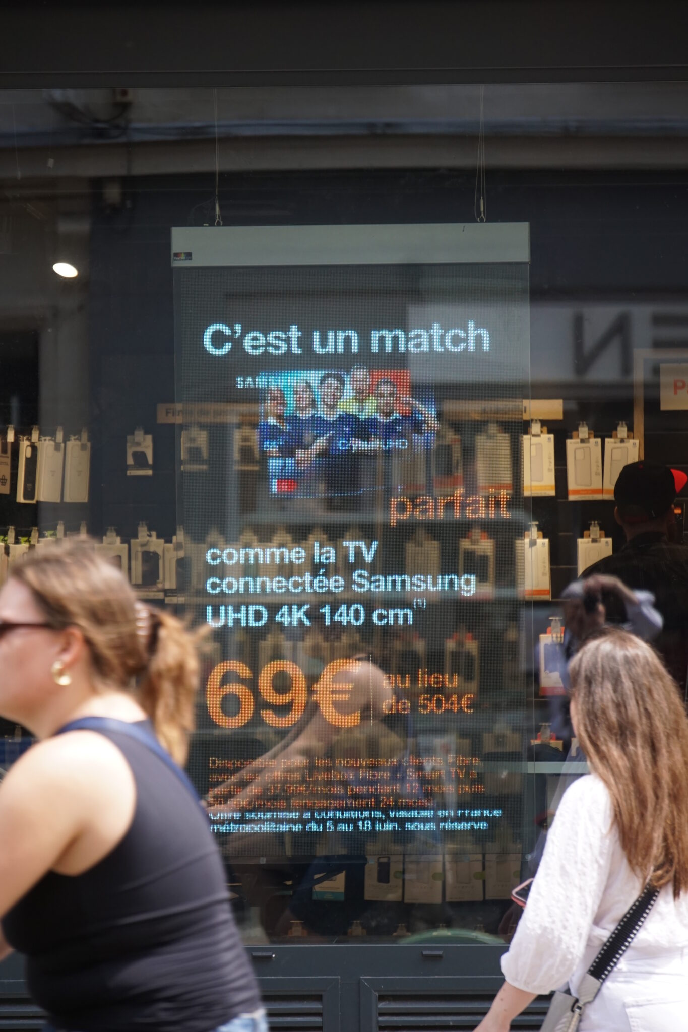 Affiche publicitaire en vitrine présentant une “TV connectée Samsung UHD 4K 140 cm” et le texte “C’est un match”, vue à travers la vitrine avec des passants en premier plan. (Note: inclut le mot-clé intégré naturellement dans la description.)