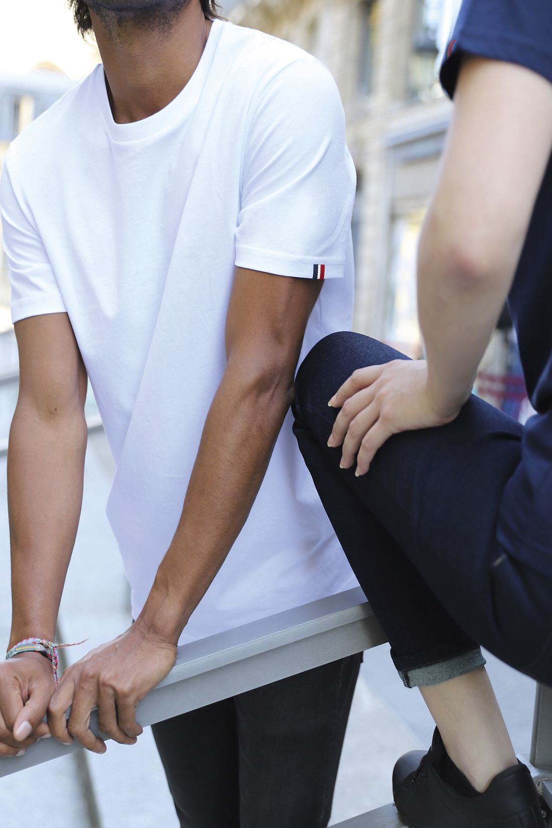 Homme en T-shirt blanc, debout dehors, les bras posés sur une rambarde; autre personne assise à côté, ambiance urbaine. Où personnaliser tee-shirt intégré naturellement.