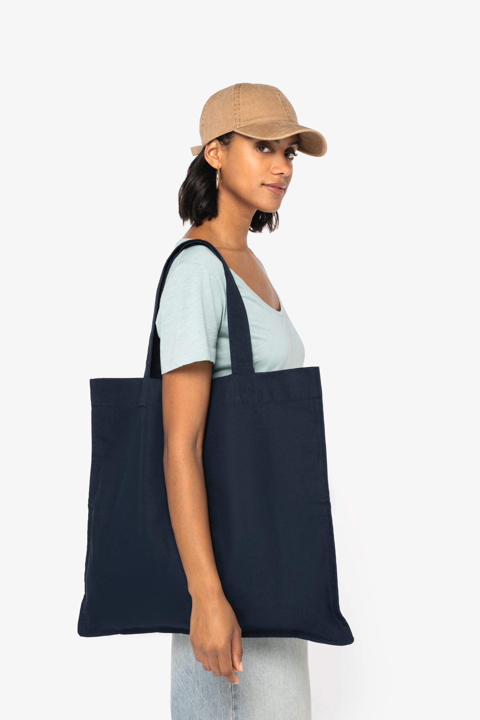 Homme ou femme portant un grand sac tote noir en coton, chemise bleu clair, casquette beige, regard latéral.