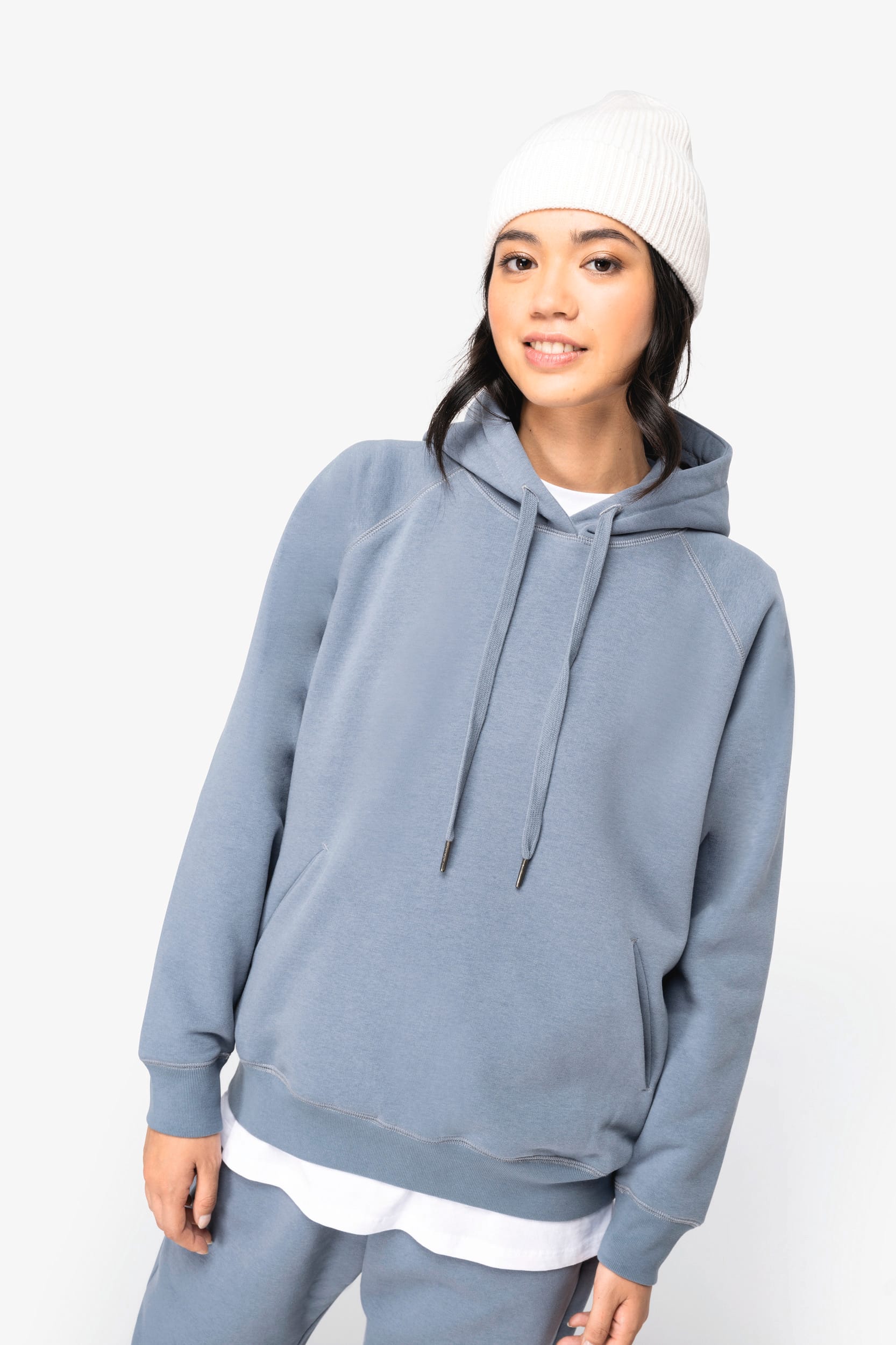 Femme portant un hoodie bleu et un bonnet blanc, debout et souriante.