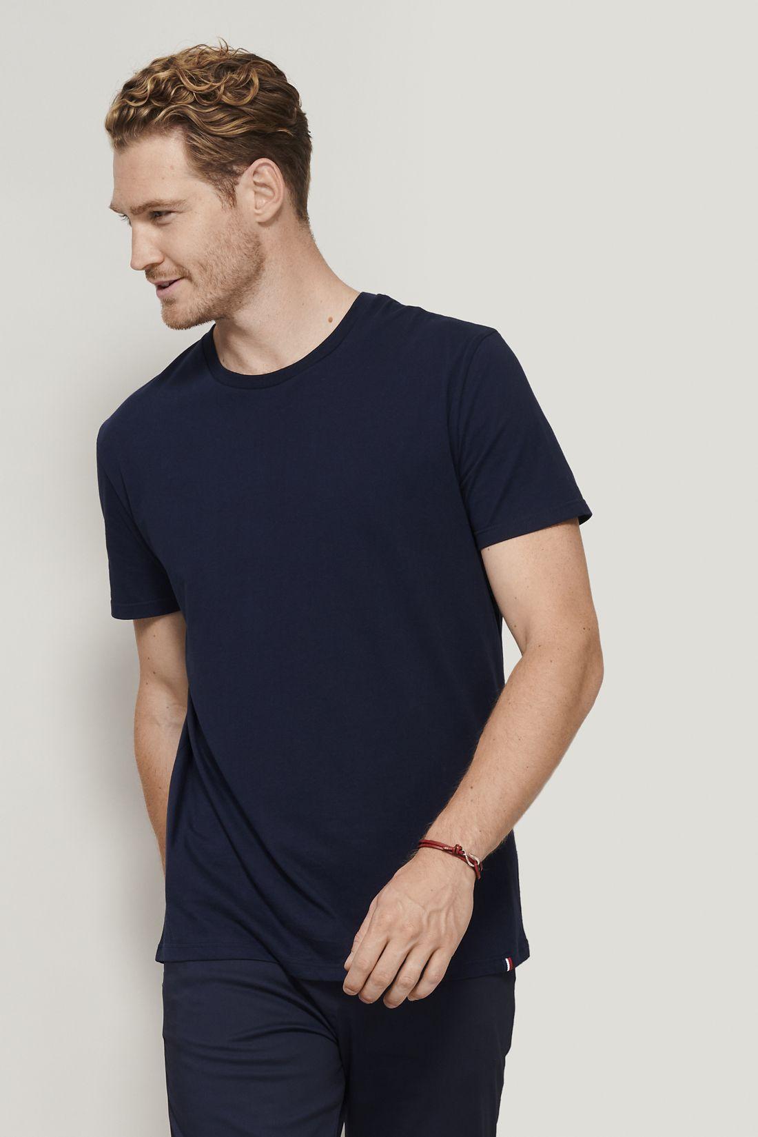 Homme portant t-shirt bleu foncé simple, coupe normale, debout contre un mur clair, regard sur la gauche. Où personnaliser tee-shirt intégré.