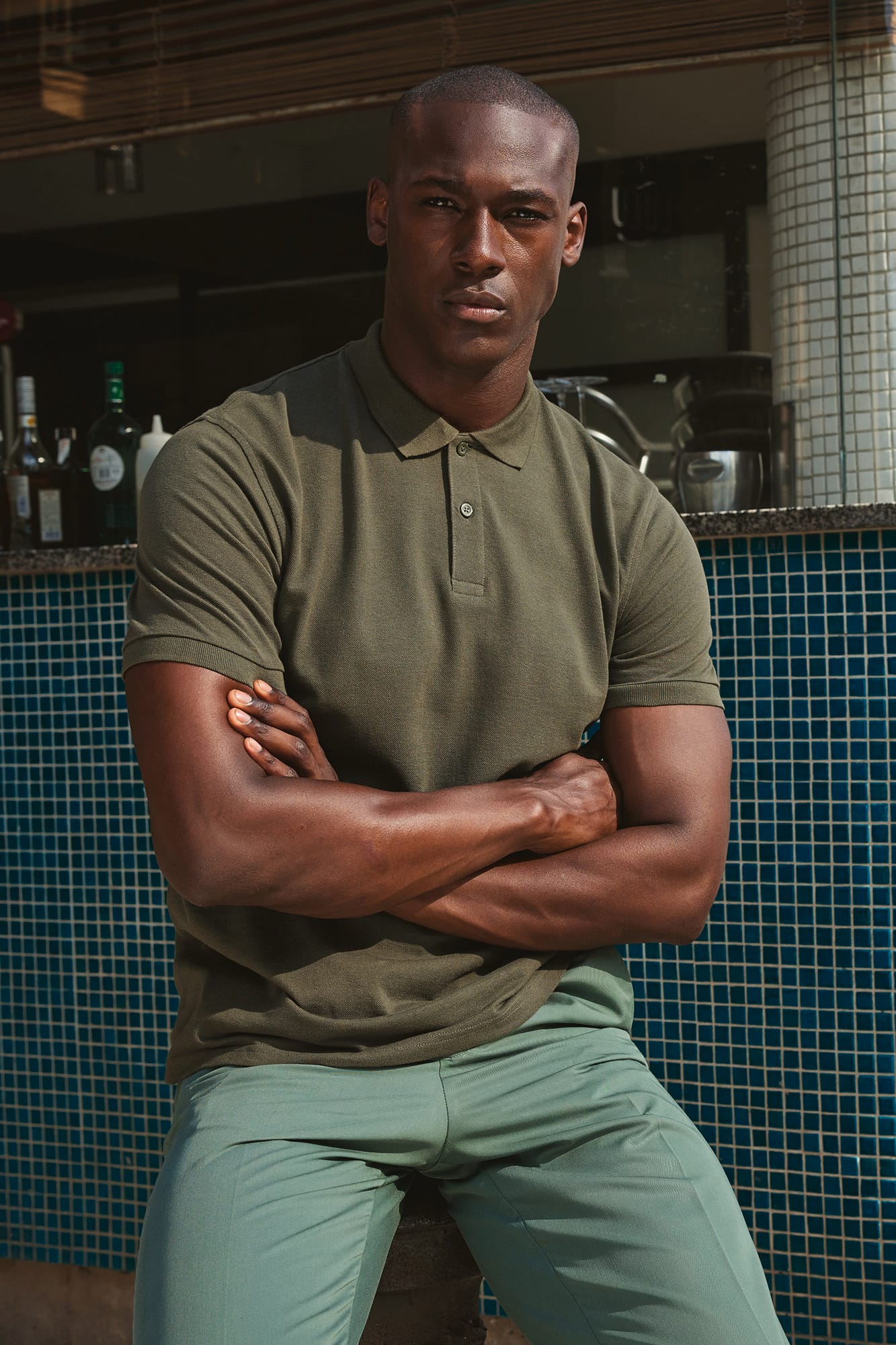 Homme portant polo olive, pose décontractée contre bar, bras croisés, tenue élégante et légère. vêtement de travail professionnel avec logo entreprise (gap).