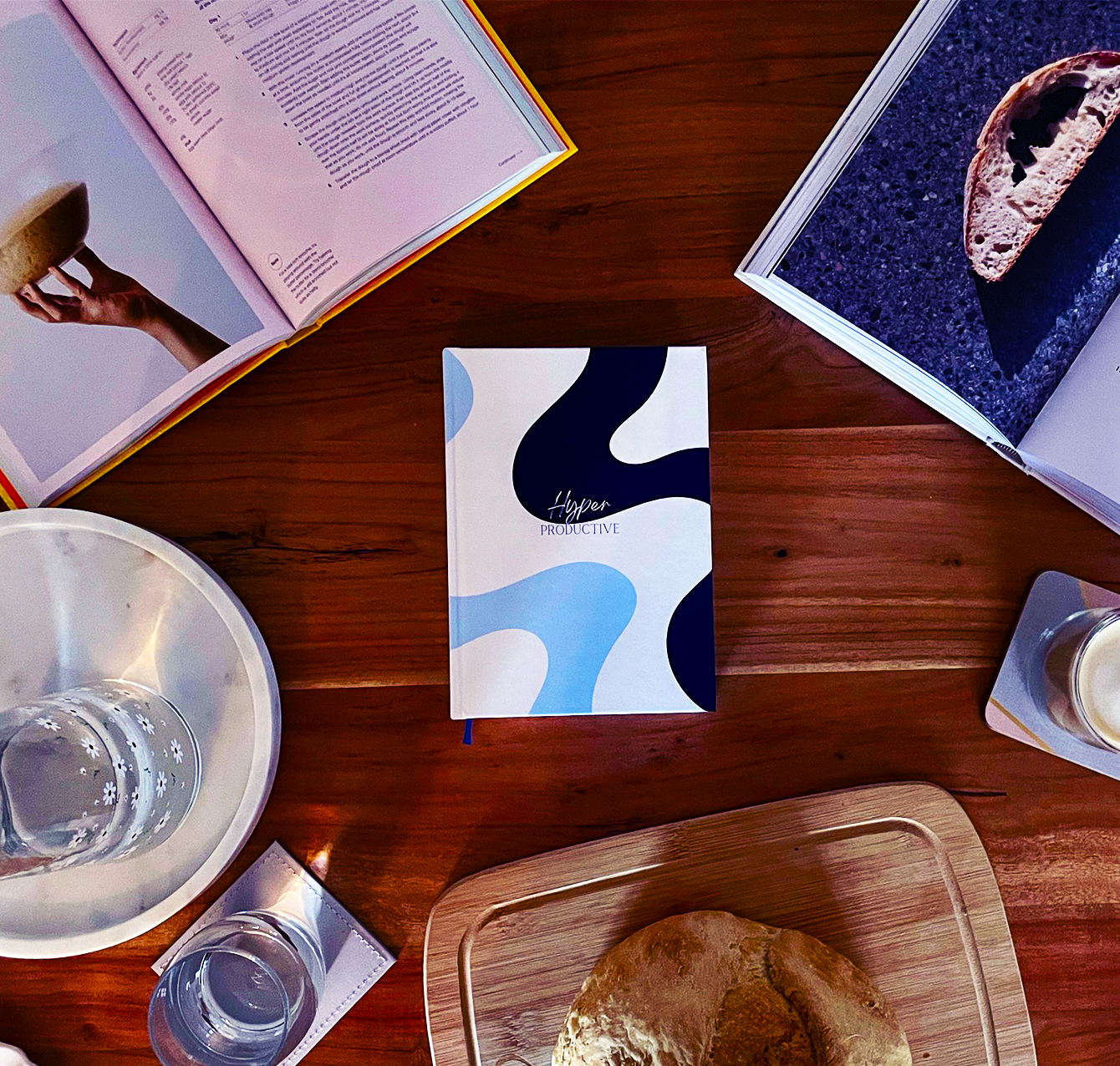 A notebook with blue and white abstract cover sits on a wooden table, surrounded by a bread roll, glass of water, open magazines, and a plate.