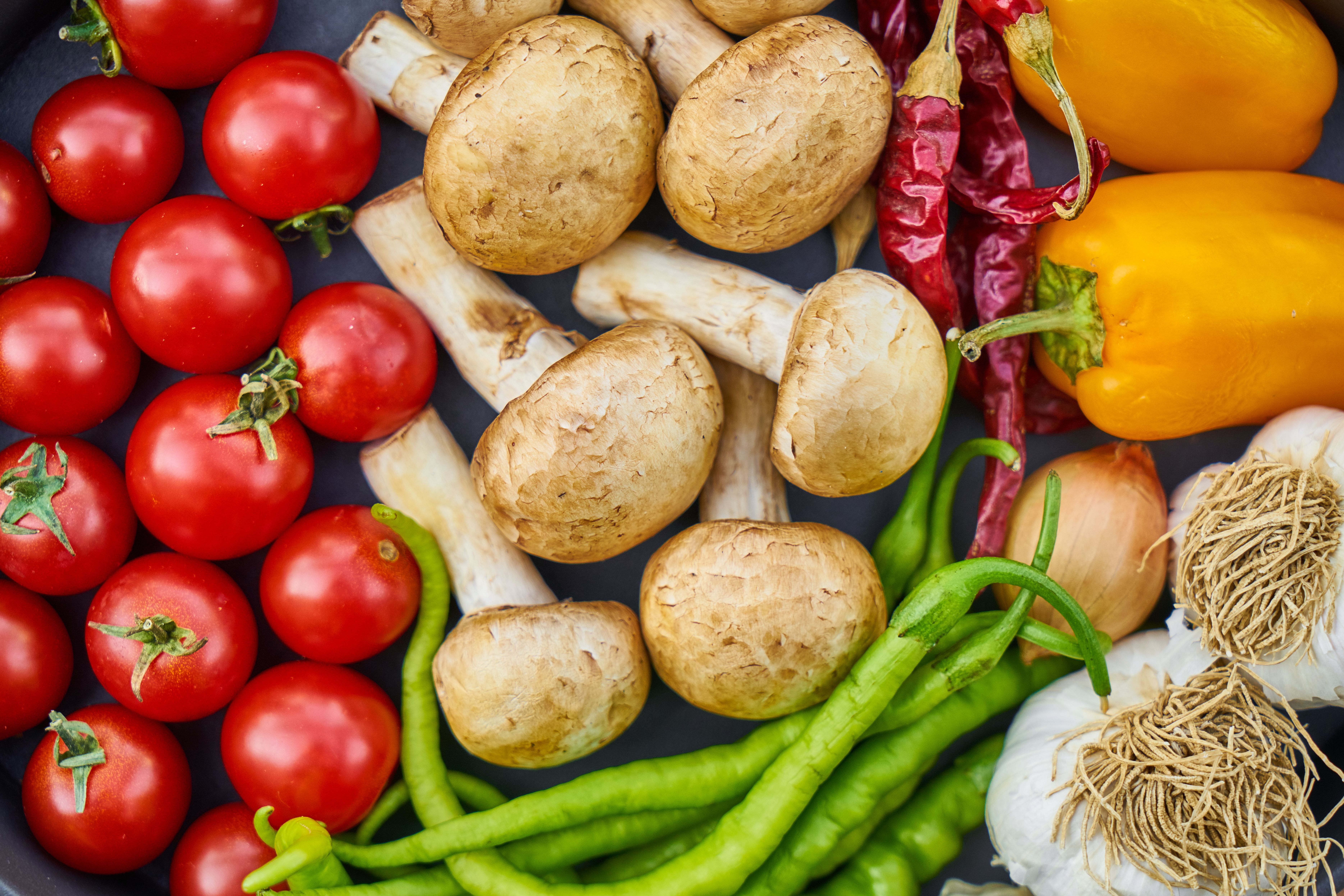 Assortiment de légumes frais, tomates cerises, champignons, poivrons jaunes, poivrons rouges, oignons et haricots verts, illustrant des produits alimentaires issus des plantes.