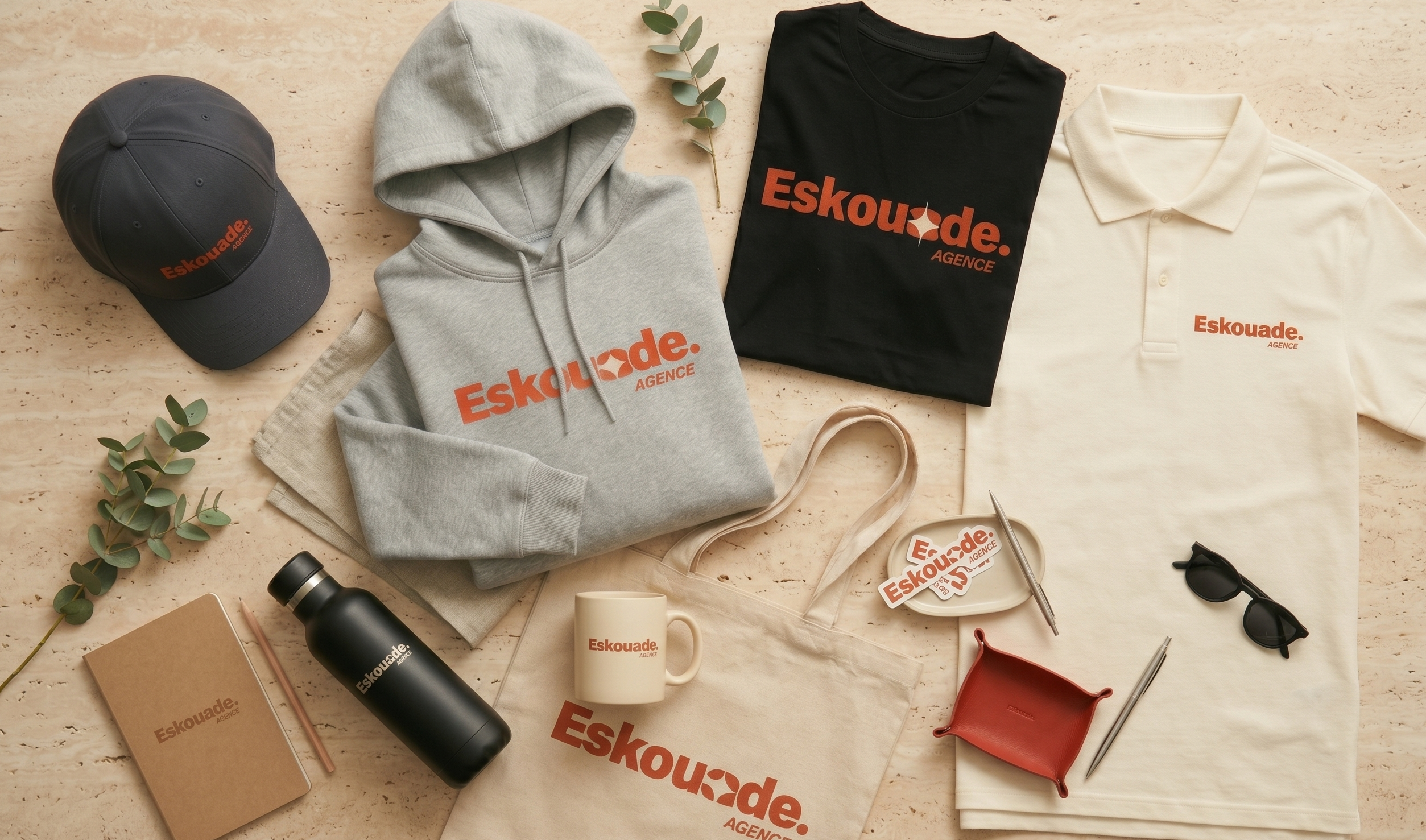 Vente de tenues d’entreprise Eskouade: casquette, hoodie gris, t-shirt et polo avec logo orange, sacs et accessoires assortis. Altier personnalisé broderie sur tablier? The image shows branded apparel. Include keyword naturally. Provide alt text: "Gamme Eskouade : casquette, hoodie gris, t-shirts et polo imprimés logo orange, accessoires assortis." But must integrate keyword: "tablier personnalisé broderie" if possible—image lacks tablier. So avoid forcing. Provide description of visible items with brand. Include keyword only if natural. It may not fit; better omit. Let's craft: "Ensemble d’articles Eskouade : casquette, hoodie gris, t-shirts et polo imprimés logo orange, sacs et accessoires assortis." 120 chars? Let's count ~120. It's fine. French. Output only alt text. Do. No quotes. Include "Eskouade" branding. Okay. Also main keyword not present; we should not force. We'll omit the keyword. But instruction says integrate main keyword if provided. It is provided. We should try include naturally. Could say "tenue de travail avec tablier personnalisé broderie" but image not tablier. So avoid misrepresentation. So omit keyword. Provide safe alt text. Let's output.