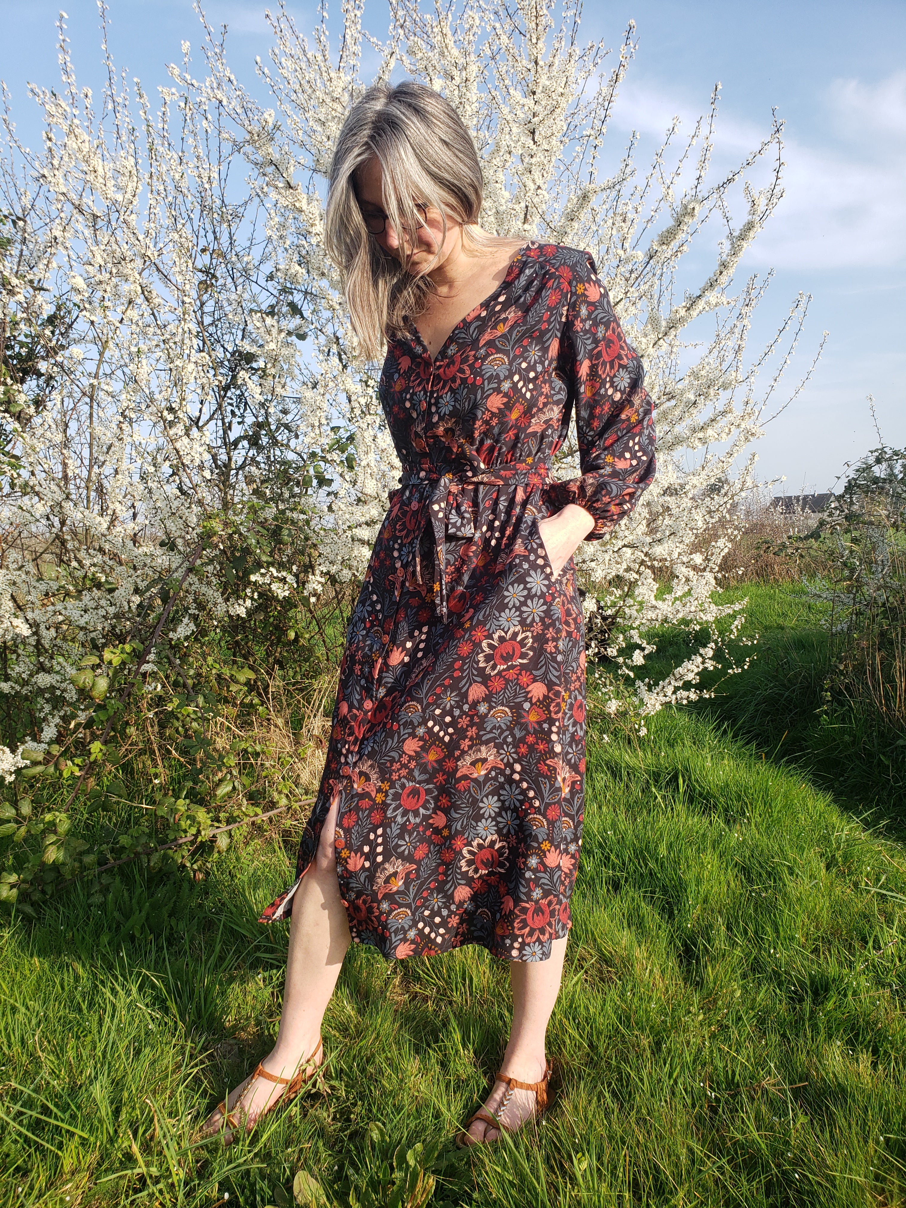 Femme portant une robe fleurie à fleurs rouges et bordeaux, ceinture à nouer, debout sur l’herbe près d’un arbre en fleurs, pieds nus dans des sandales. Apprendre la couture de a à z intégré naturellement si possible? but alt text: include keyword naturally. Let's craft: "Femme en robe longue imprimée florale, ceinture à nouer, debout dans un jardin en fleurs, idéal pour apprendre la couture de a à z." But must describe visible; we should include keyword integrated. Ensure not awkward. I'll write: "Femme en robe longue à imprimé floral, ceinture nouée, debout dans un jardin en fleurs, illustration pour apprendre la couture de a à z." Keep 125 chars. Let's count approx. We'll output. No quotes. Also ensure no extraneous. Let's finalize.