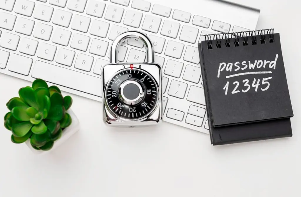 Cadenas à combinaison posé sur un clavier d'ordinateur blanc à côté d'un carnet noir affichant 'password 12345', illustrant la nécessité d'un coffre-fort de mots de passe.