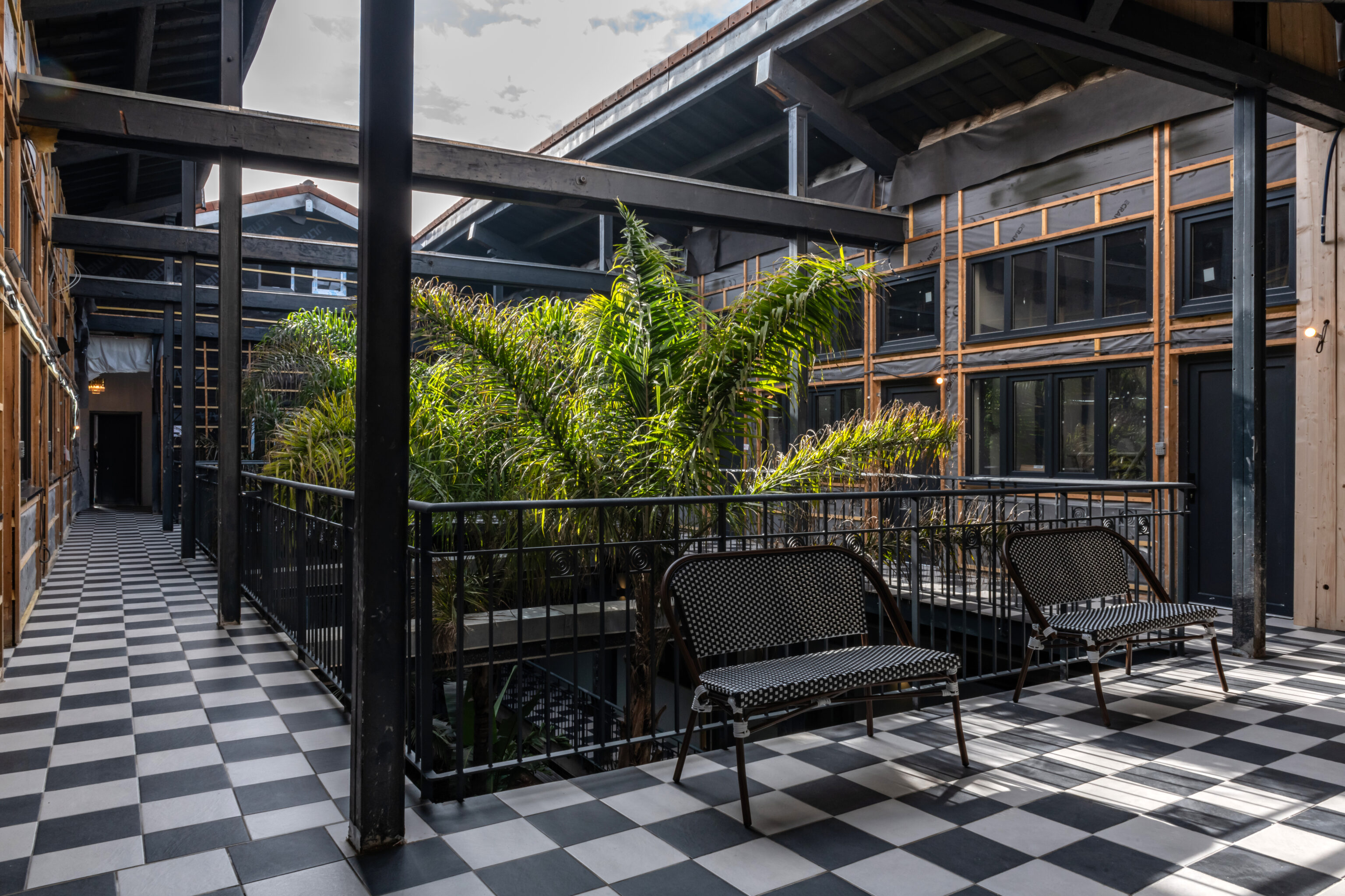 Couloir en mezzanine d’un bâtiment moderne avec véranda et plantes vertes, bancs noirs en osier et sol à damier noir et blanc. Luminosité naturelle présente. Luminosité idéale d'un écran intégrée.