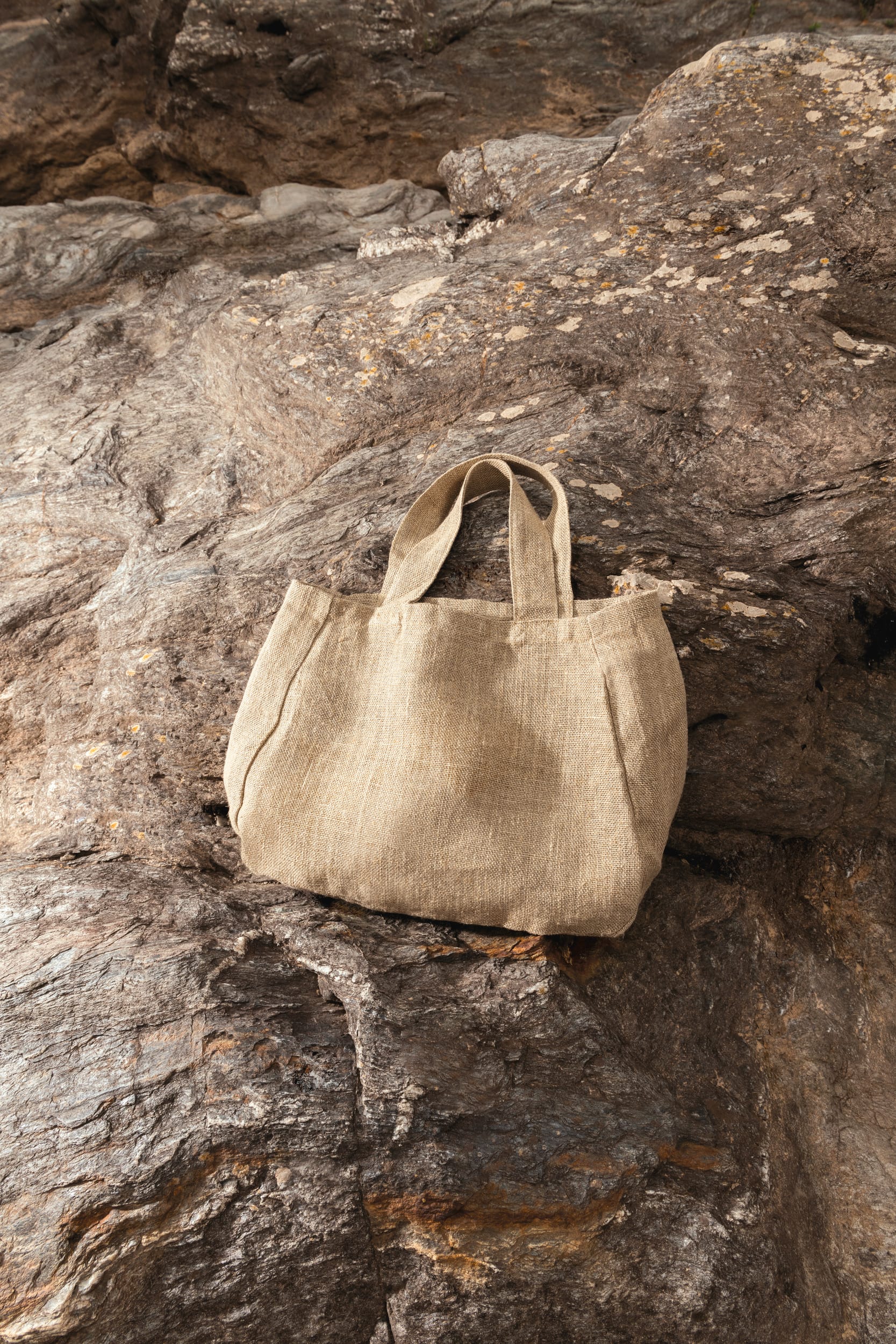 Petit tote bag en lin beige posé contre une paroi rocheuse, avec poignées simples et pochettes latérales apparentes. Intègrez le choix d'un tote bag.