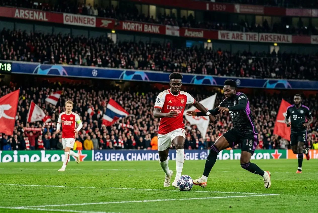Joueur d’Arsenal en blanc et rouge contrôlant le ballon face à un adversaire en noir, Premier plan sur le terrain et une affiche UEFA Champions League en arrière-plan. pronostic ligue des champions 2026 intégré naturellement.