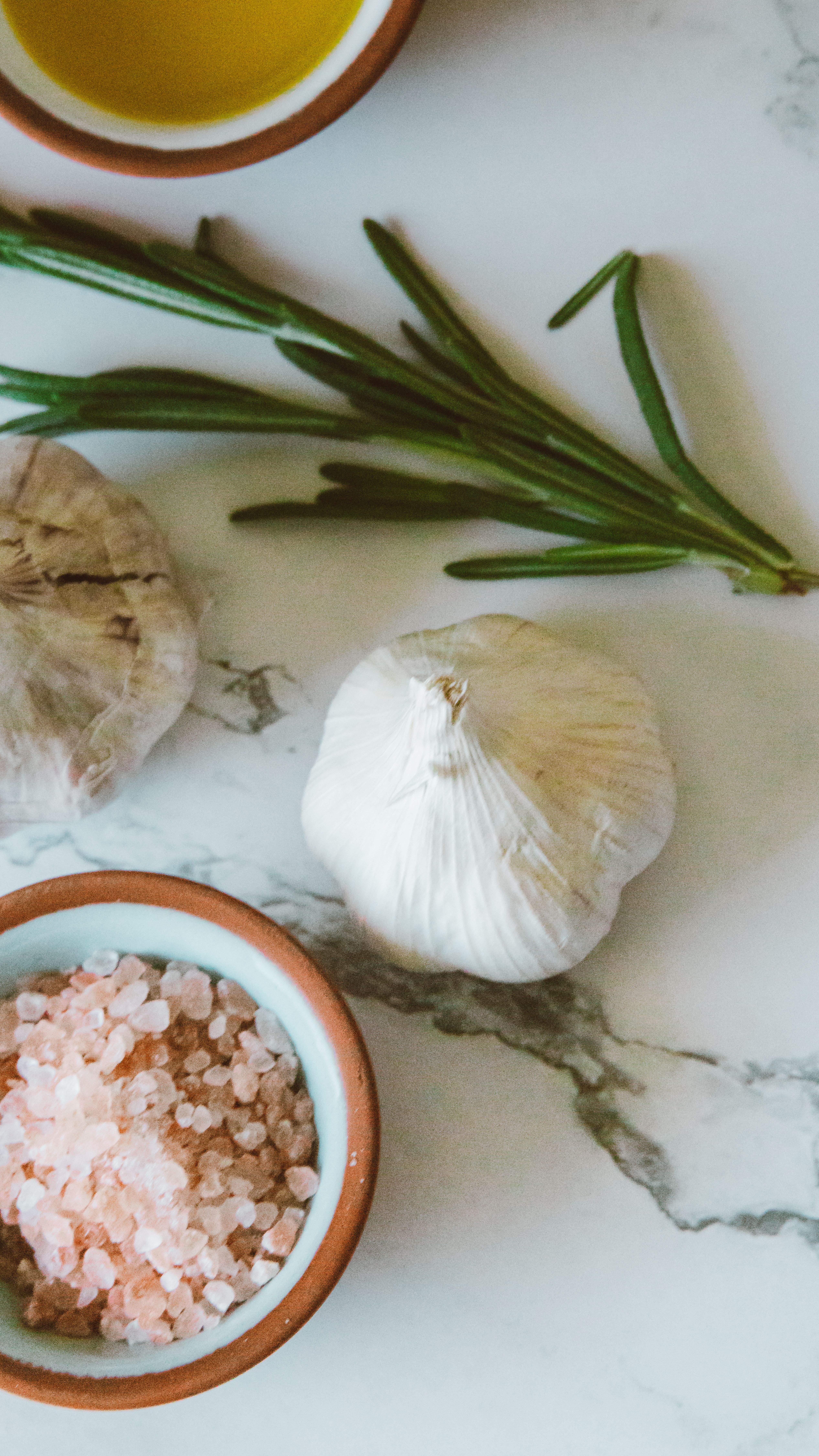 Herbs and a garlic clove near a bowl of pink salt on a marble background.