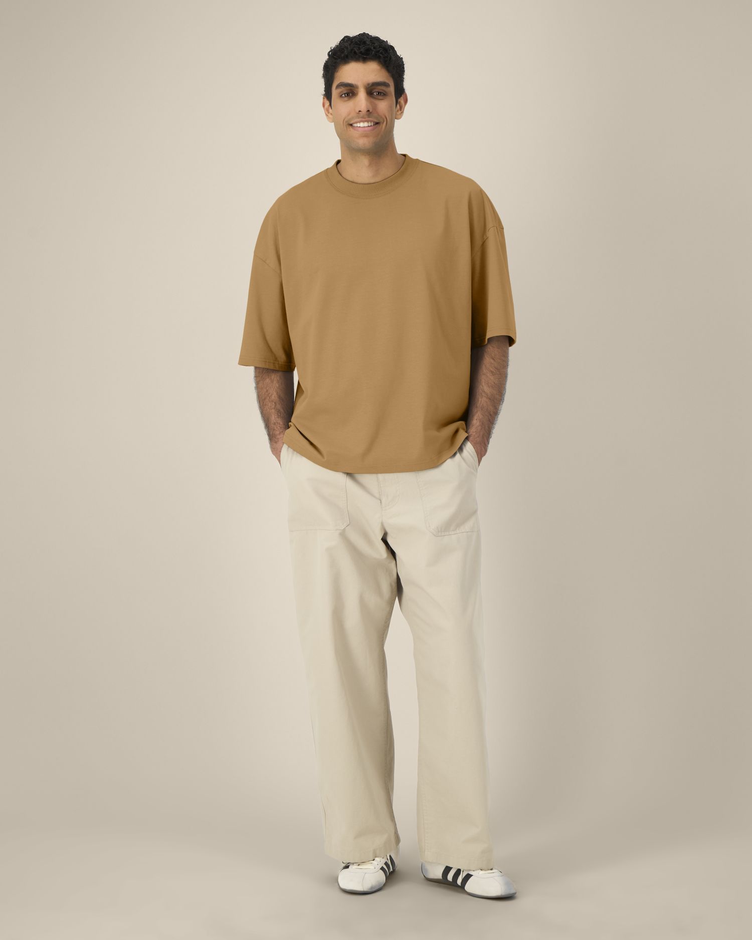 Un homme debout porte un T-shirt beige ample et des pantalons cargo blancs, les mains dans les poches.
