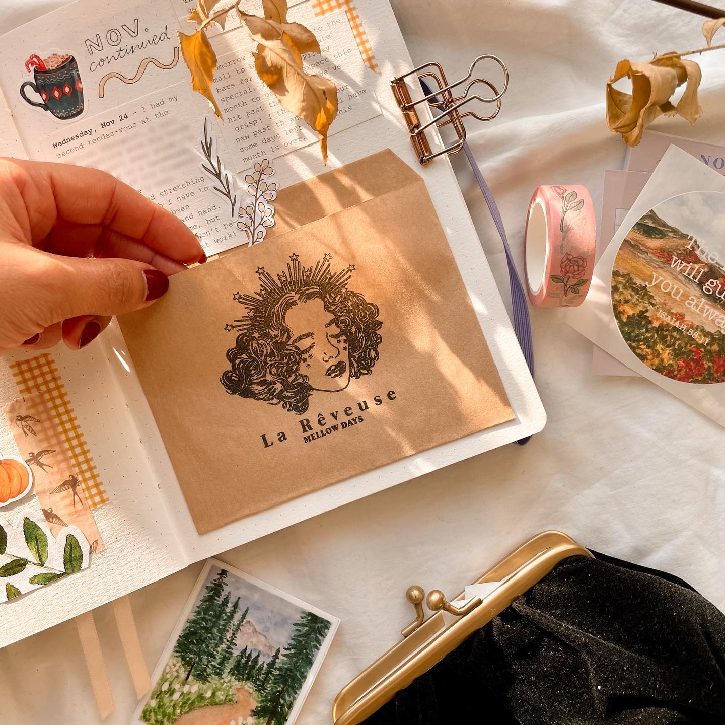 Brown paper envelope with a vintage illustration of a woman's face and crown, labeled “La Révéeuse MELLOW DAYS” on a desk with journaling supplies. Brown paper envelope with a vintage illustration of a woman's face and crown, labeled “La Révéeuse MELLOW DAYS” on a desk with journaling supplies.