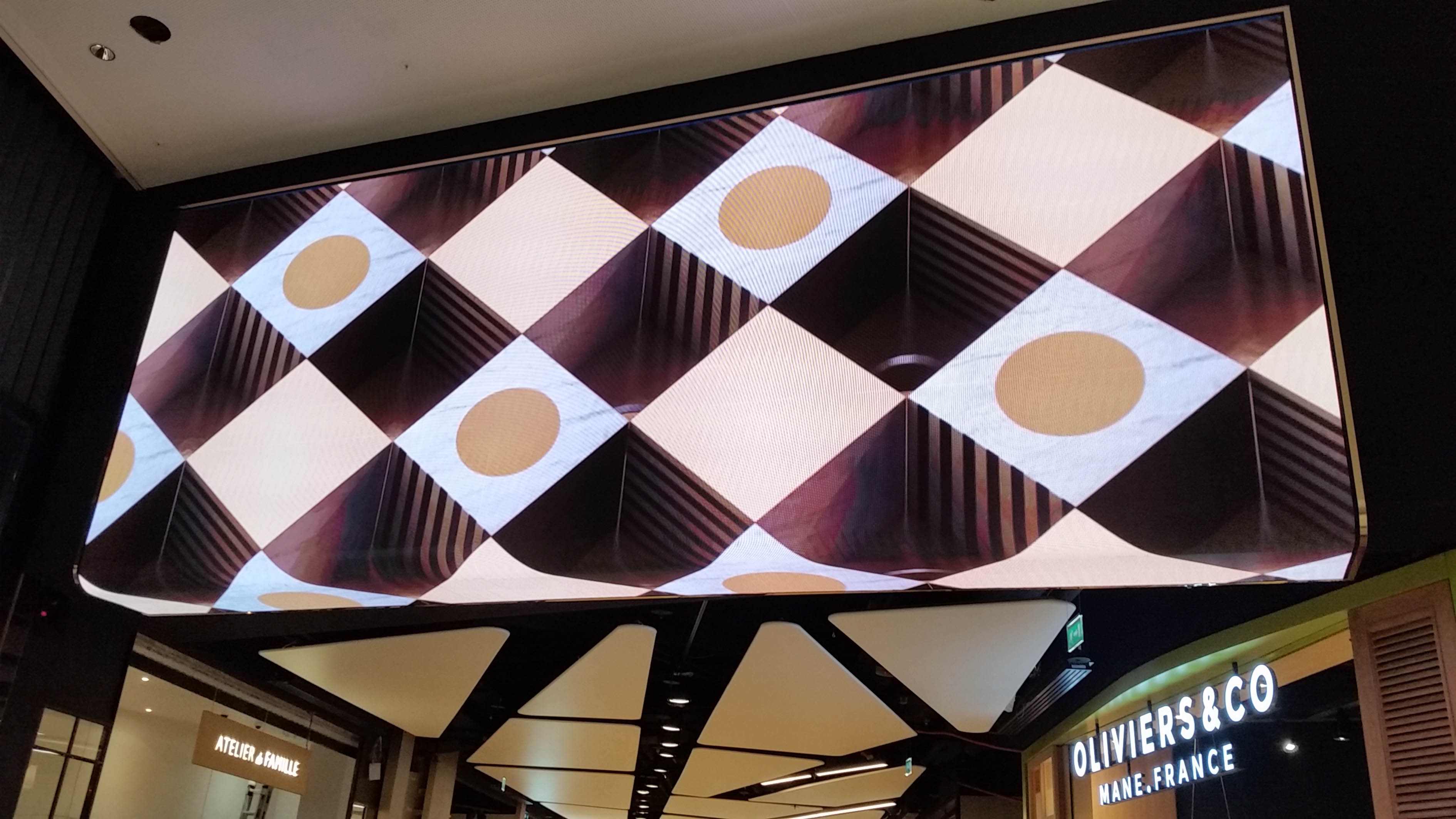 Affiche géante ou écran publicitaire incurvé montrant un motif alterné de carrés noirs et blancs avec cercles dorés, suspendue dans un centre commercial. intégré motif motif écran lcd? pas nécessaire. Include keyword: "probleme affichage écran lcd" naturellement: "Affichage présentant un problème affichage écran lcd sur une grande vitrine publicitaire". But need description of visible image; include main subject. Let's craft: "Écran publicitaire géant affichant un motif en damier avec cercles dorés, suspendu dans un centre commercial." Then include keyword integrated: "problème d’affichage écran lcd" hmm must be natural: "Écran publicitaire géant présentant un problème d’affichage écran lcd, suspendu dans un centre commercial." That's acceptable. 118 chars approx. Let's finalize.