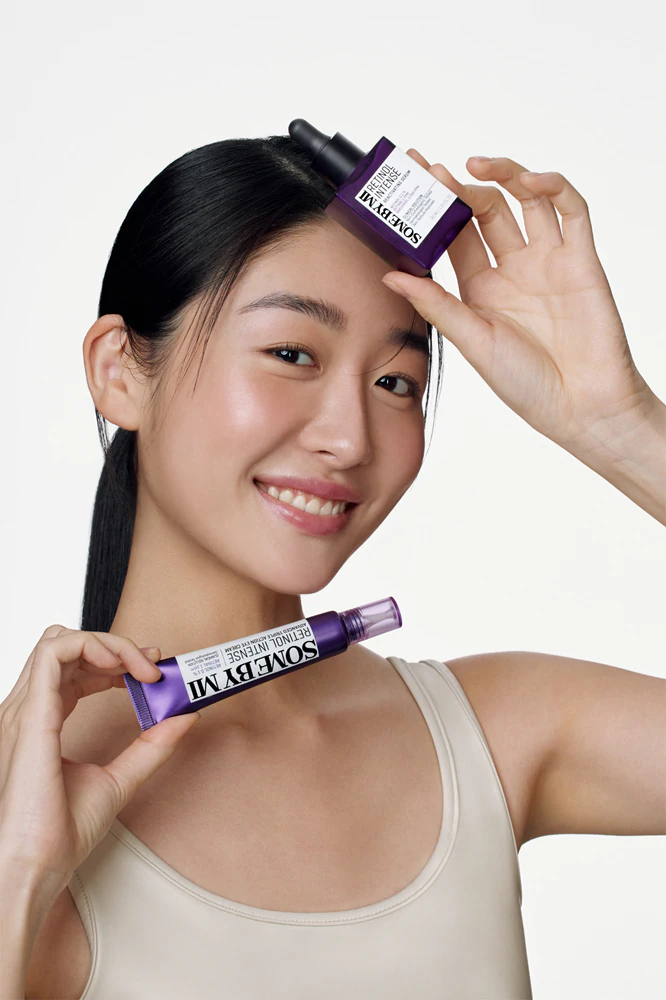 Jeune femme asiatique souriante tenant deux tubes de serum Sombre Moon, l’un près du front et l’autre près du menton, sur fond clair.