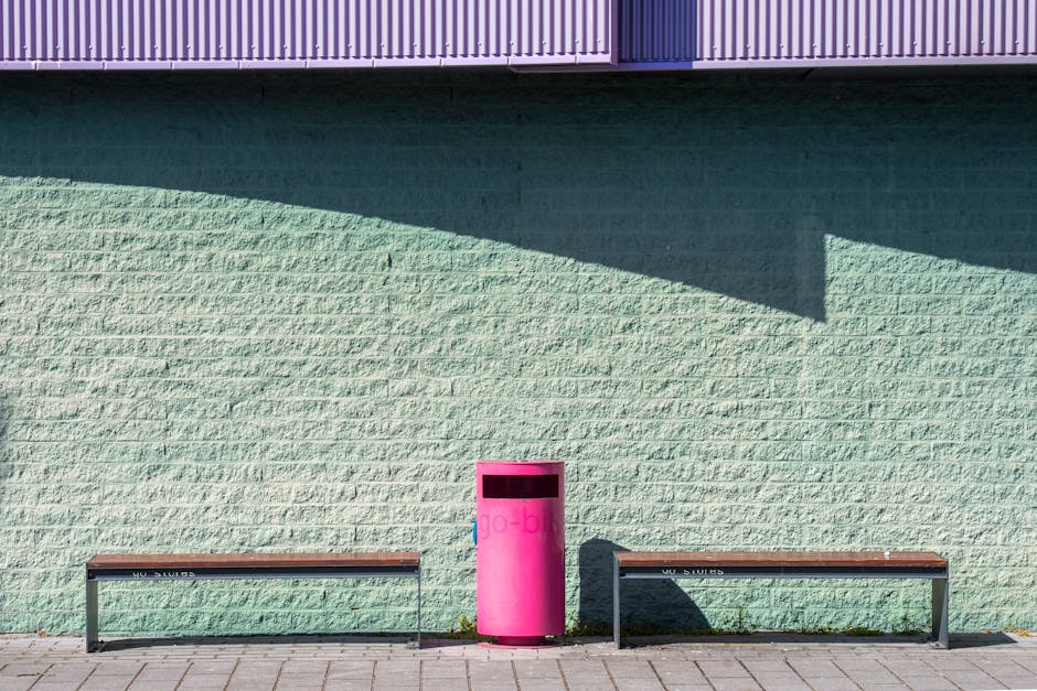 Une scène urbaine minimaliste avec des bancs et une poubelle rose vif contre un mur coloré.