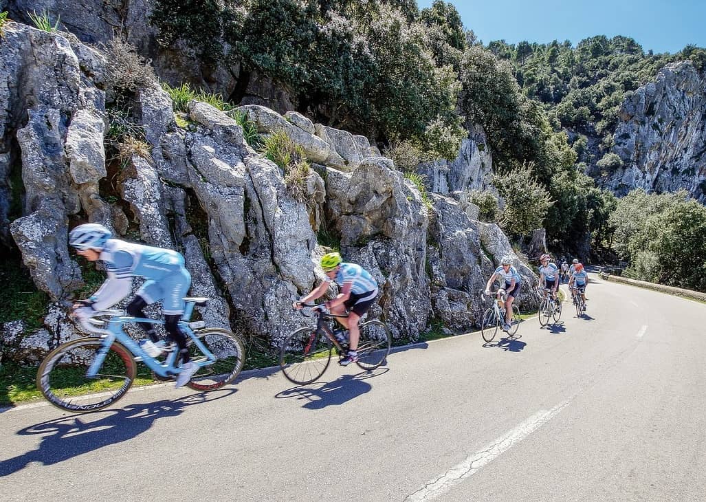 Groupe de cyclistes en descente sur une route côtière entourée de rochers, en plein air, avec casque et équipements de vélo. Vélo route Majorque en contexte touristique.