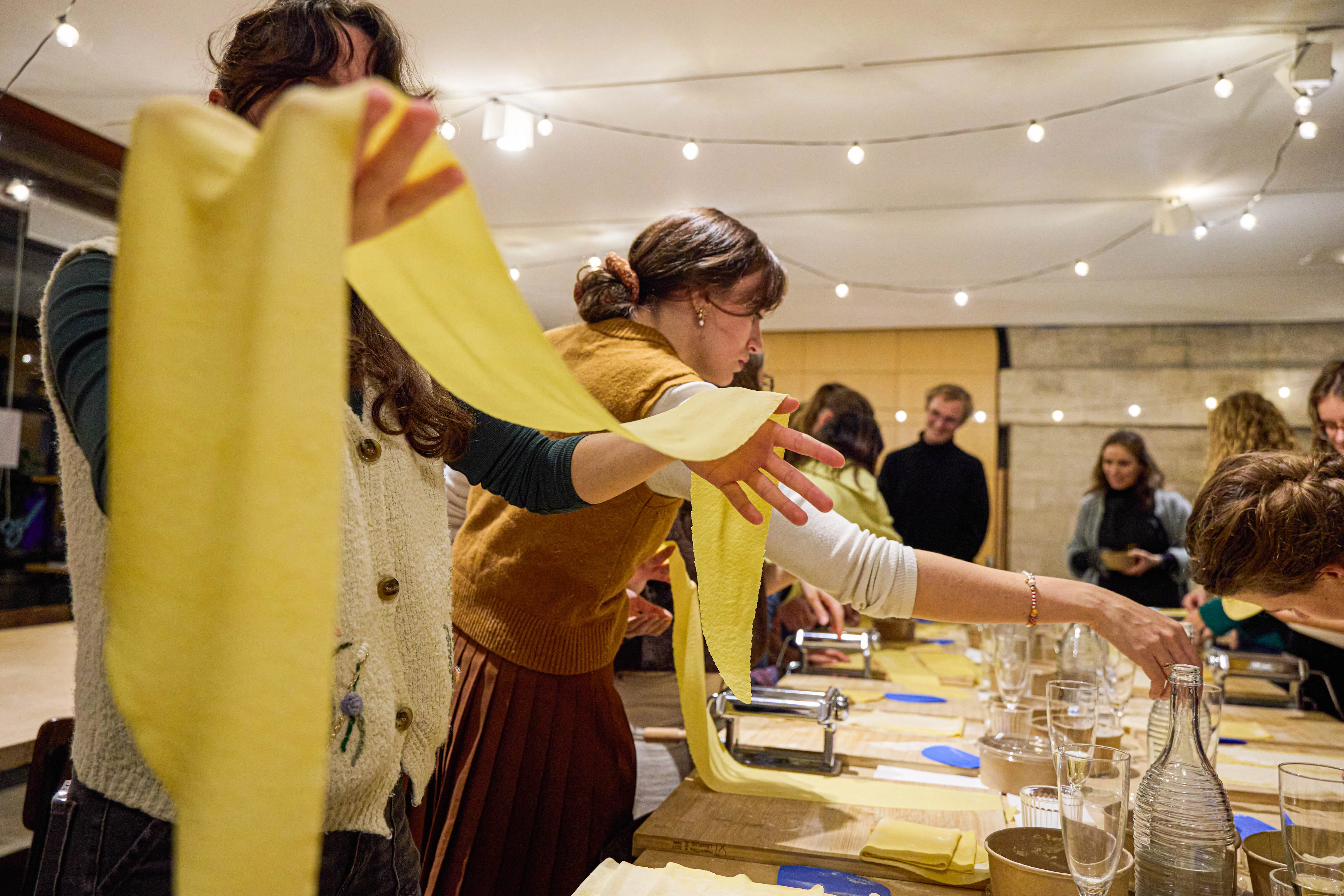 Participants en atelier cuisine italienne, déployant des longes de pâte fraîche sur des machines à pâtes, ambiance conviviale et lumineuse.