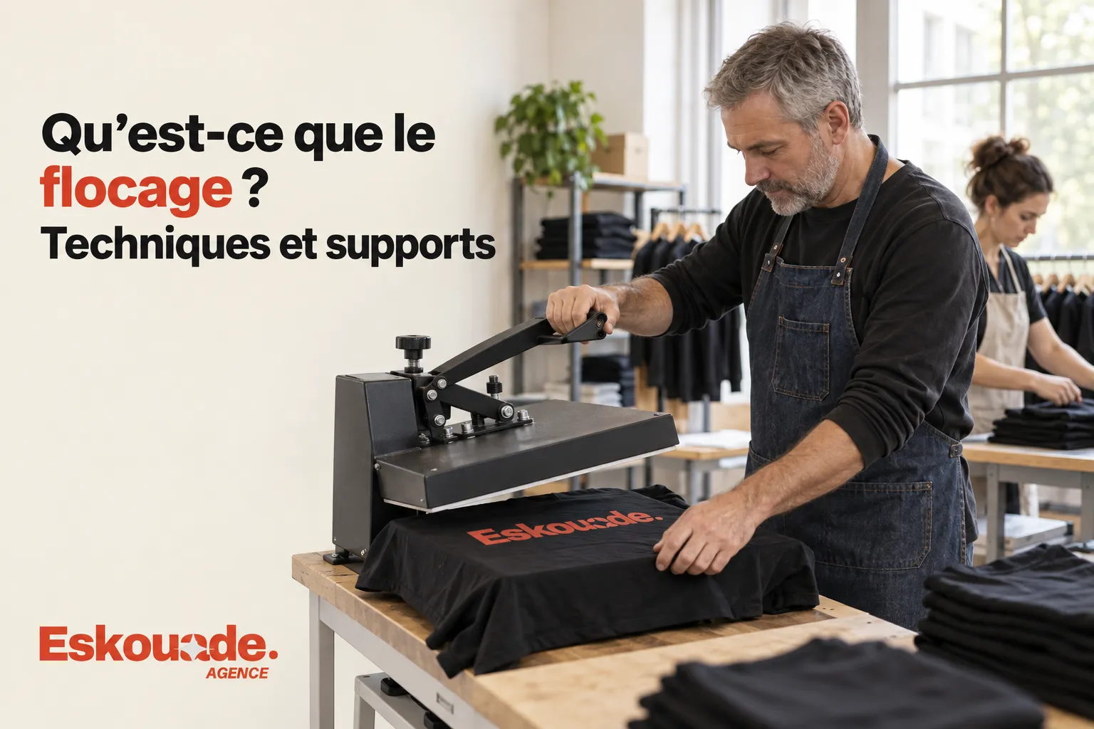 Un homme portant un tablier presse un t-shirt noir sur une presse à flocage dans un atelier.