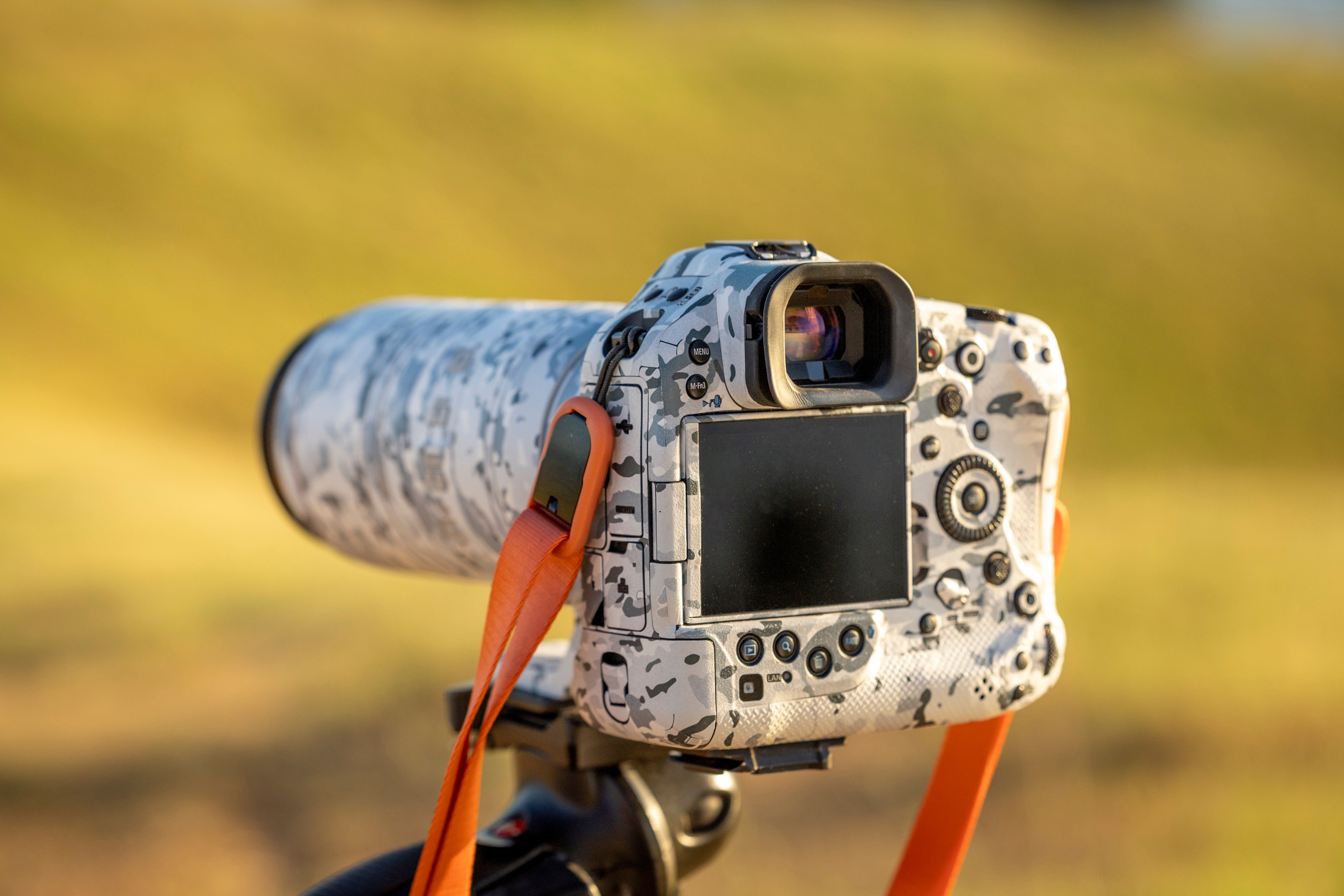 Caméra reflex argentée avec objectif long et capteur face arrière, sur trépied, en extérieur, style camouflage, boutonnières orange. objectif pour photo animalière est suggéré.
