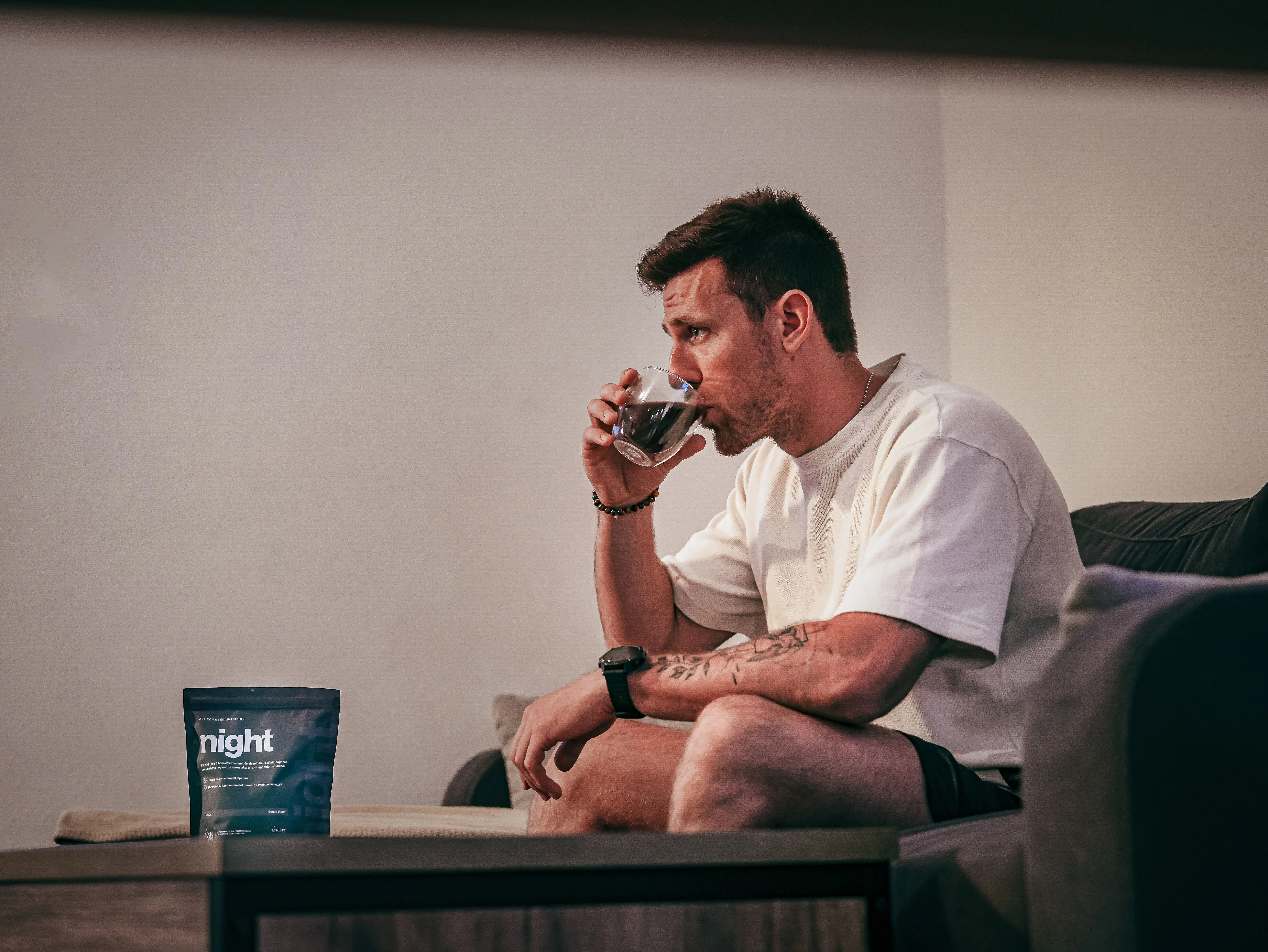 Homme assis sur un canapé, boit un verre et regarde droit devant; à côté, sachet bleu nuit "night" posé sur la table.