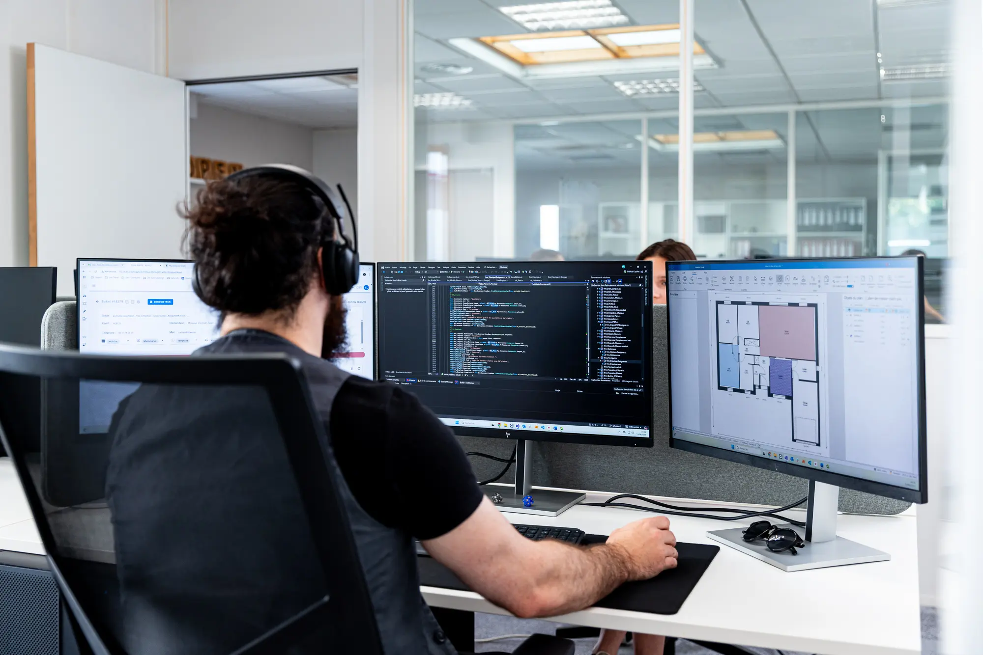 Développeur devant trois écrans affichant du code et des plans, en posture de travail dans un bureau. Développeur devant trois écrans affichant du code et des plans, en posture de travail dans un bureau.