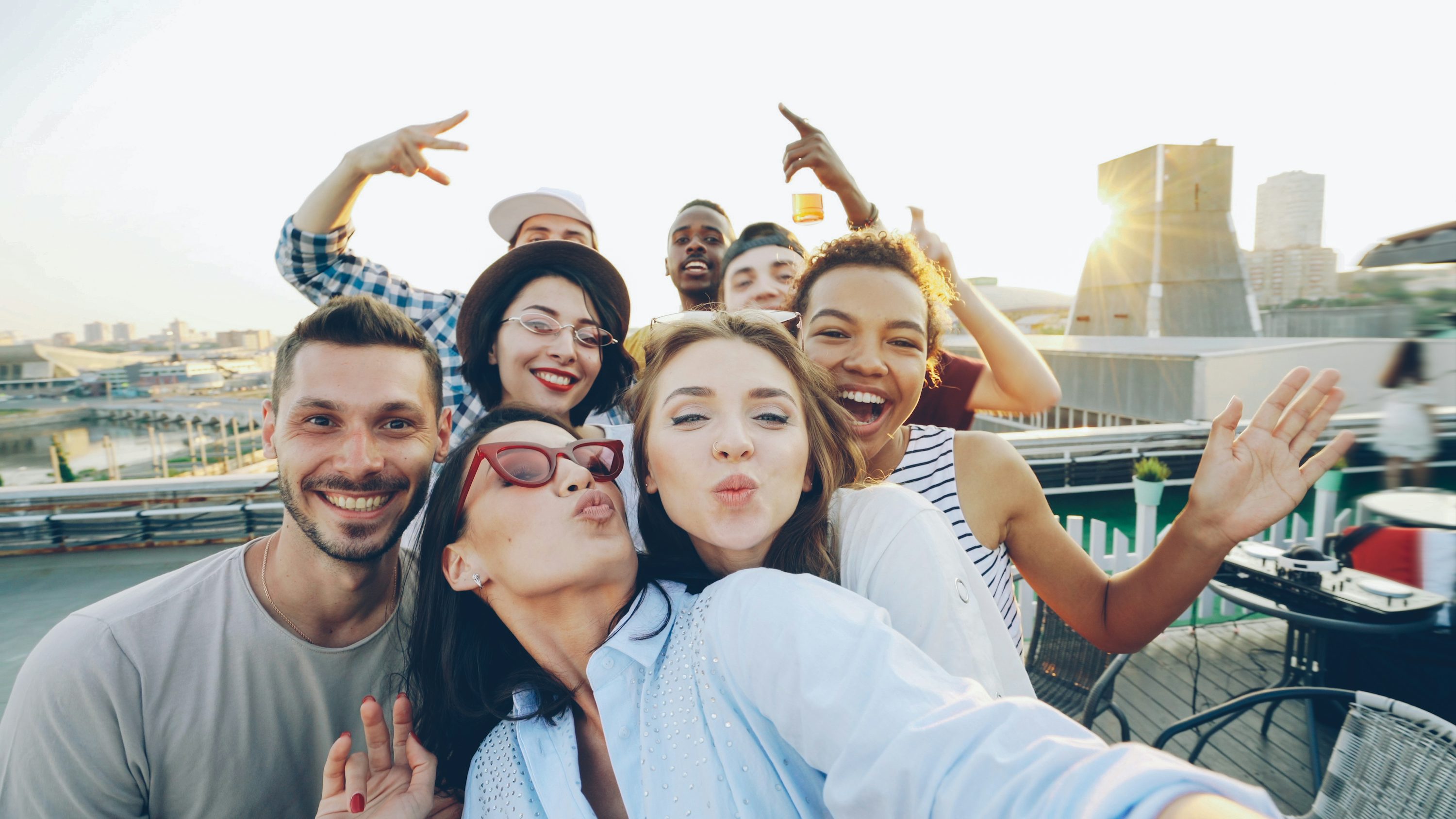 Groupe de jeunes adultes prenant un selfie au coucher du soleil sur une terrasse, sourire et gestes festifs.