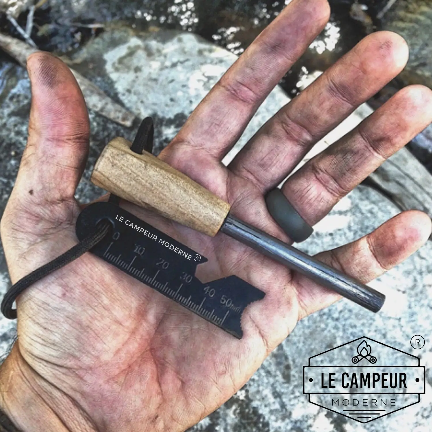 Laisse de poche métallique noir et manche en bois, outil de survie posé sur une paume sale; logo "Le Campeur Moderne" en bas à droite.