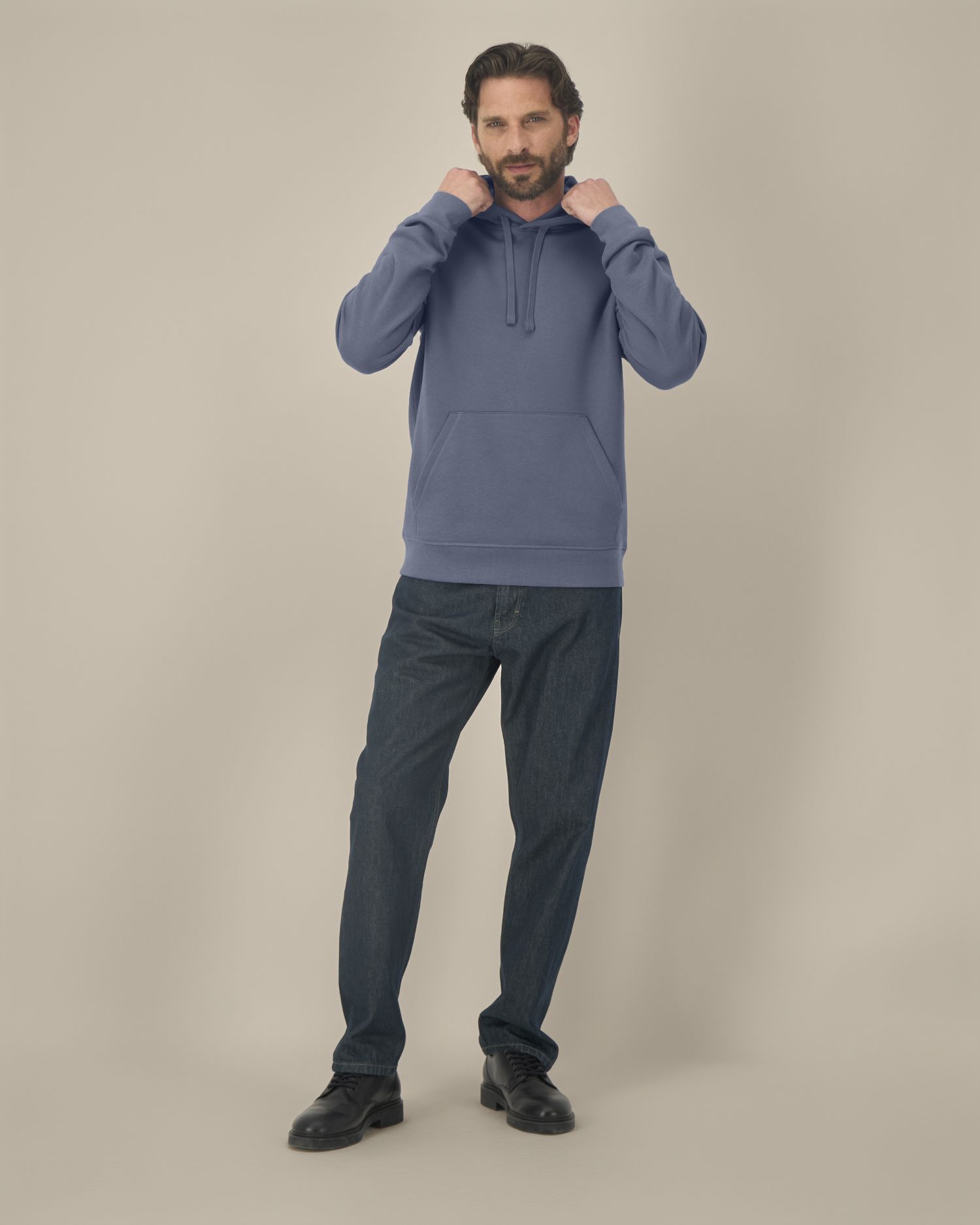 Homme debout portant sweat à capuche bleu et jeans, pose décontractée contre fond beige, aligne le regard. Atelier de marquage textile france pour entreprise.