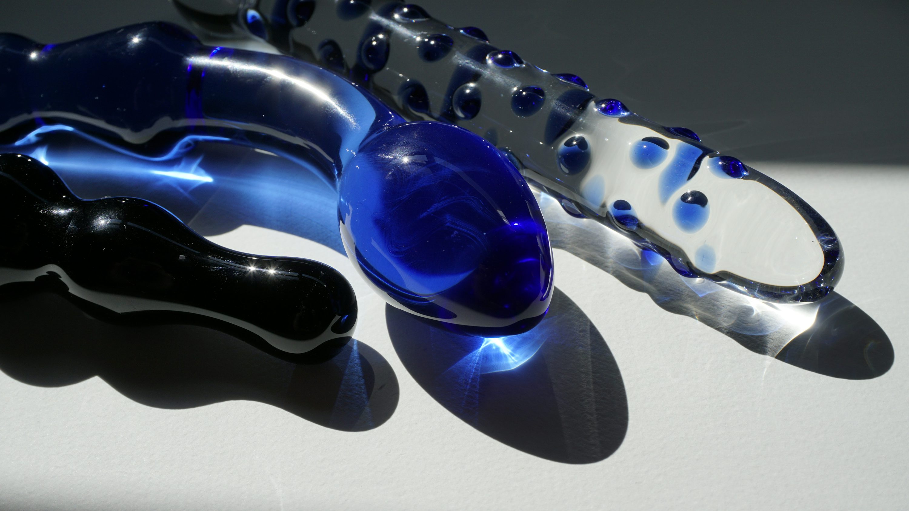 Gode en verre bleu translucide et objets similaires transparents, formes organiques, reflets et ombres sur fond clair.