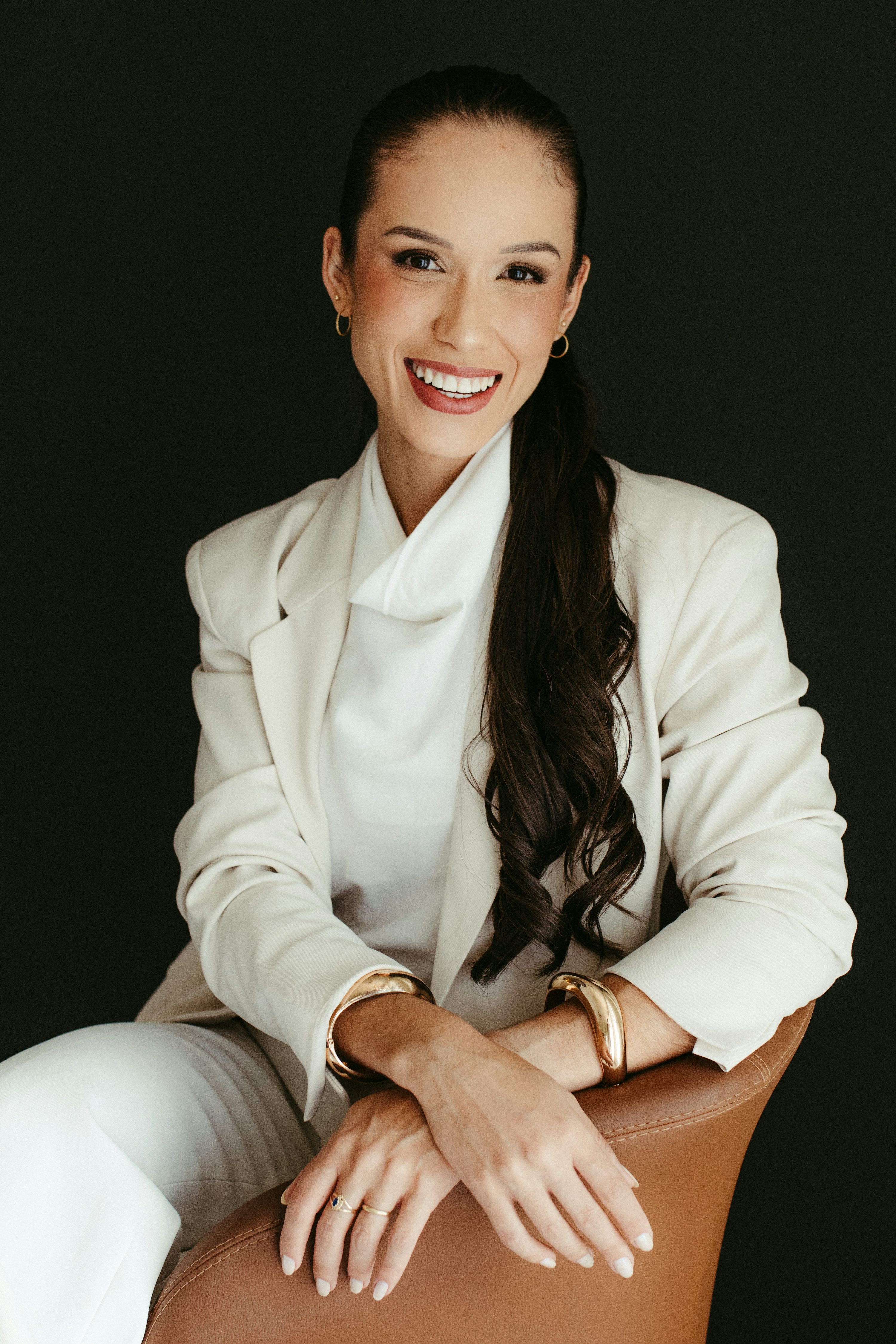 Une femme souriante en tailleur blanc pose assise devant un fond sombre.