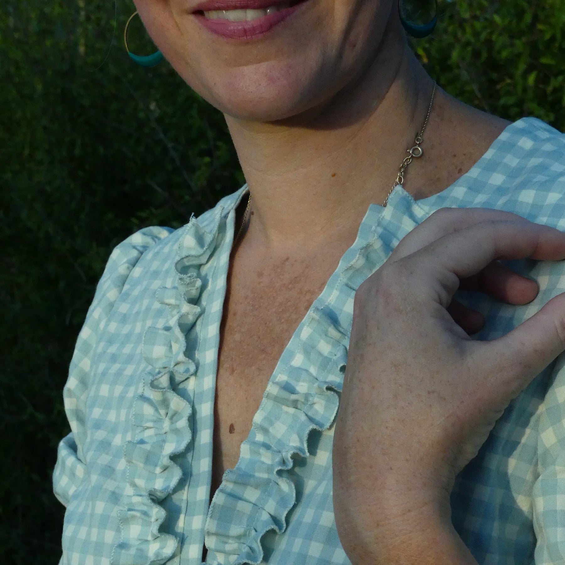 Détail d’une blouse à vichy bleu clair avec volants le long de l’encolure, porté par une personne, boucles d’oreilles et peau claire.