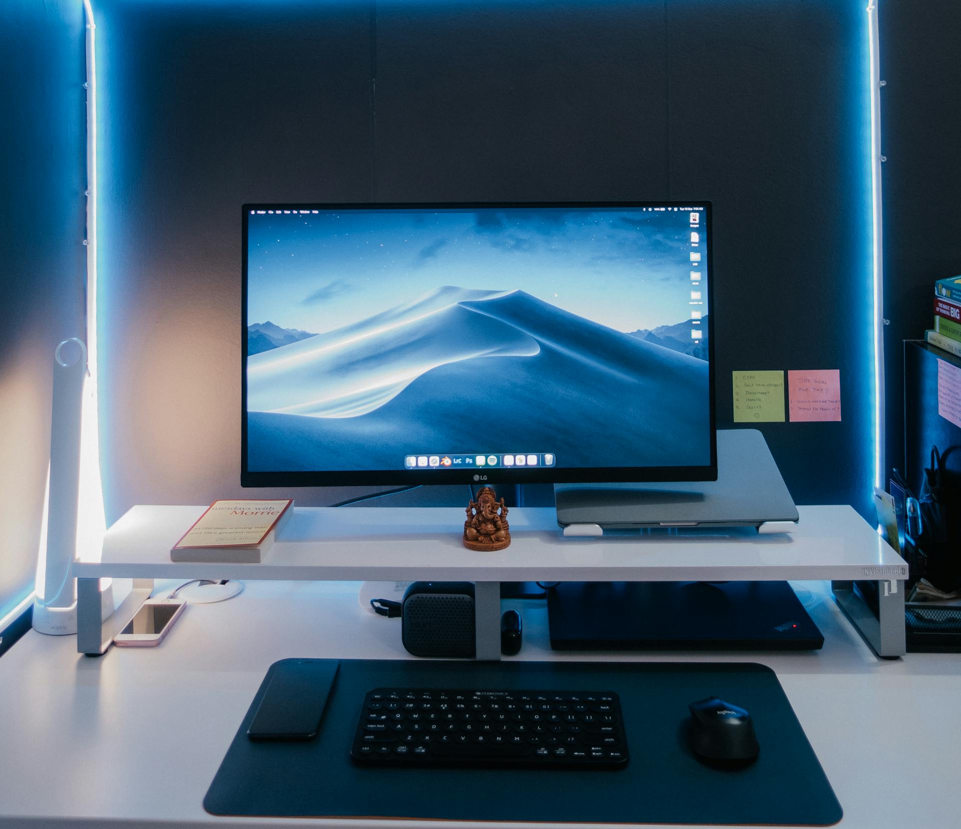 Bureau clair avec écran affichant un fond désertique, clavier et souris sur un tapis, lampe LED et objets décoratifs sur le bureau. Meilleur ventilateur chambre silencieux.