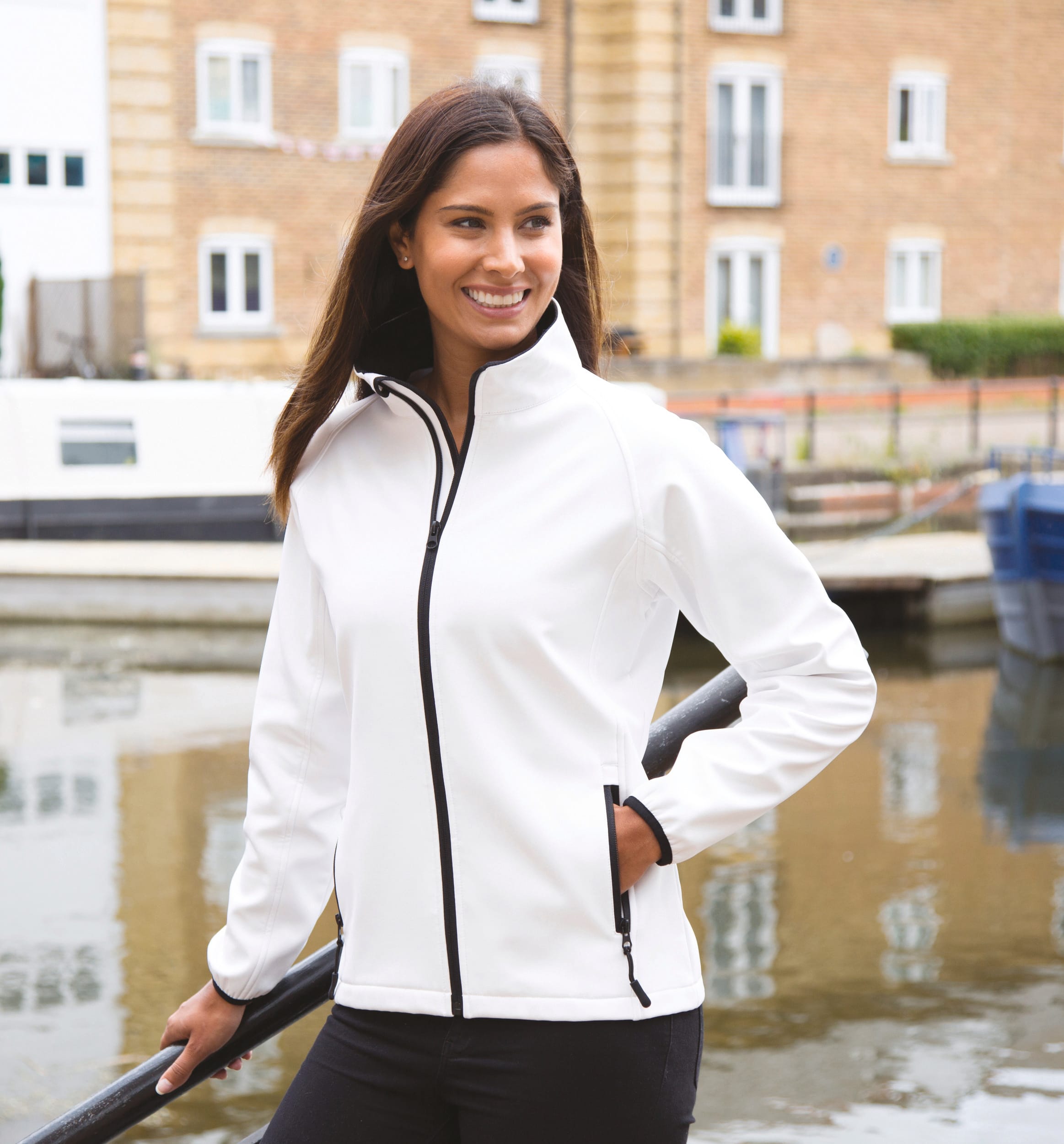 Veste polaire blanche zippée avec poches, femme souriante près d’un canal urbain, détail noir sur les fermetures et poches. entretien de polaire personnalisée brodée intégré.