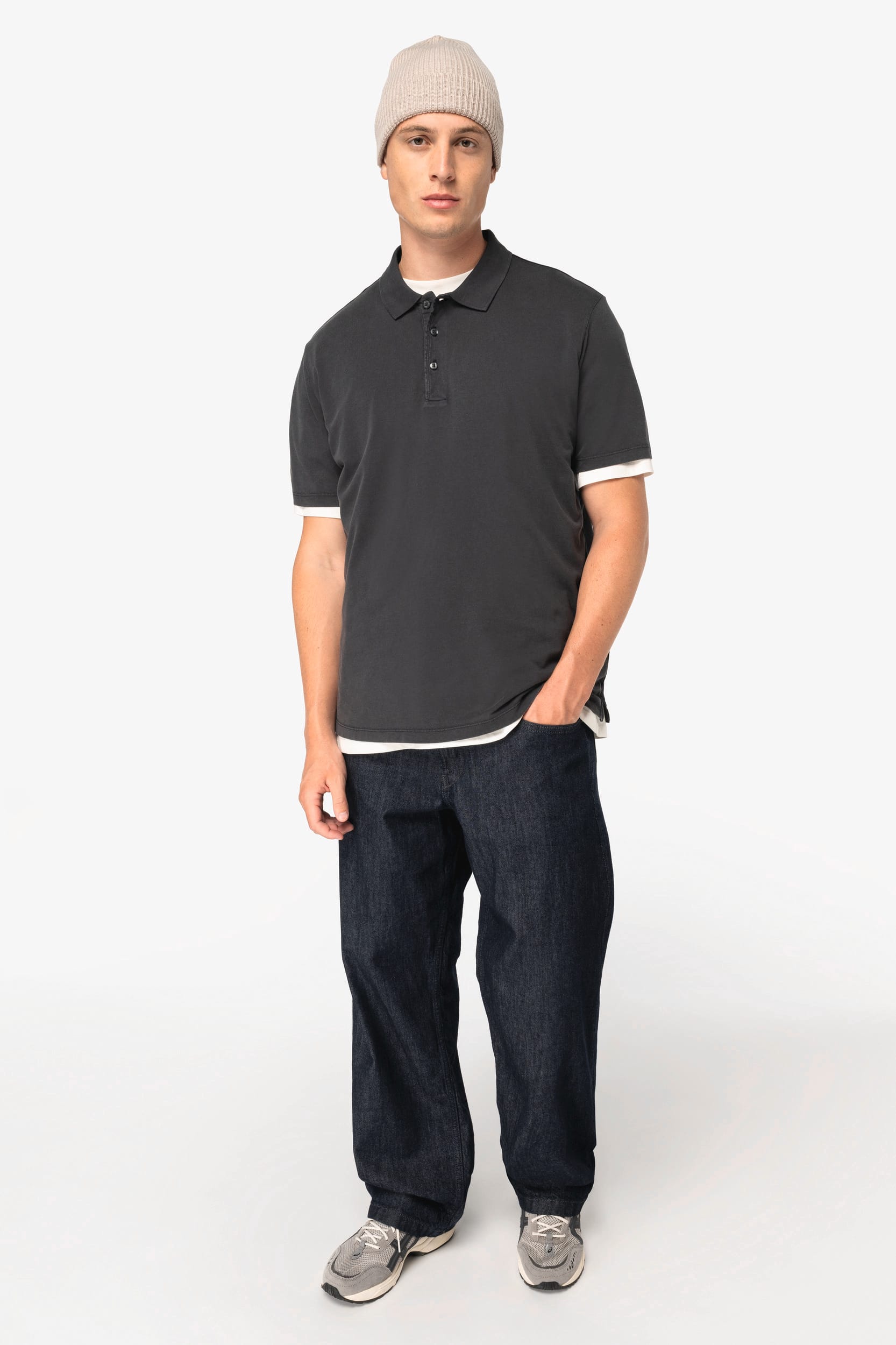 Homme portant polo gris, jean sombre et bonnet beige, debout face à l’appareil photo.