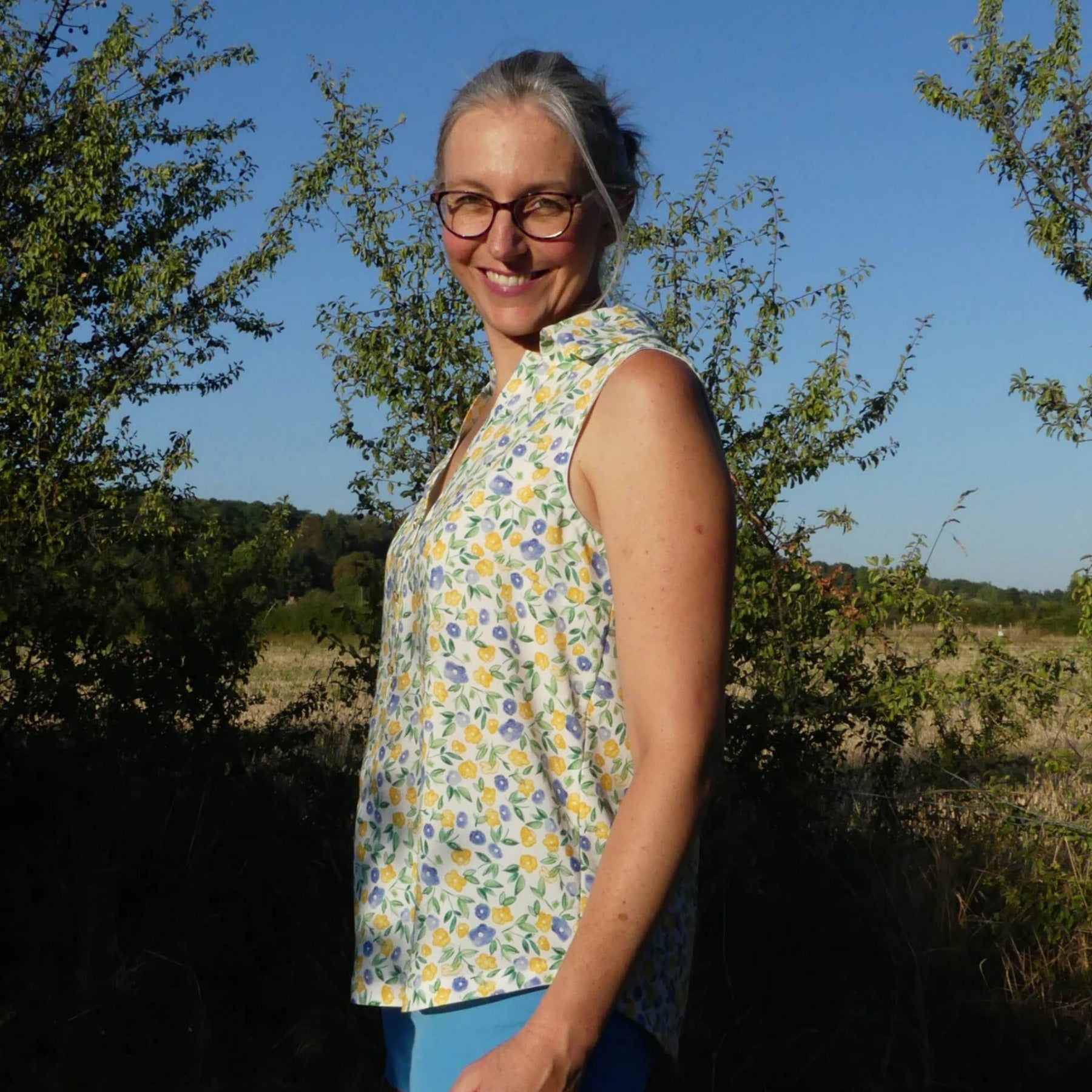 Femmes debout en plein air, étendue à la lumière du soleil, portant un top sans manches fleuri jaune et bleu, avec lunettes, sourire et cheveux gris. intégration du mot-clé: comment lire un patron de couture? Aucun contexte direct. Après vérification, inclure le mot-clé de manière naturelle dans la description: "Femme portant un top fleuri, idéale pour comprendre comment lire un patron de couture." mais must be alt text describing visible. Include keyword seamlessly. Let's craft: "Femme souriante portant un top fleuri jaune et bleu, debout en plein air, prête à apprendre comment lire un patron de couture." Ensure length 125 chars. Count: "Femme souriante portant un top fleuri jaune et bleu, debout en plein air, prête à apprendre comment lire un patron de couture." That's long. Let's shorten: "Femme souriante en haut fleuri jaune et bleu, debout en plein air, prête à comprendre comment lire un patron de couture." That's okay. Characters ~125. Output. Femme souriante en haut fleuri jaune et bleu, debout en plein air, prête à comprendre comment lire un patron de couture.