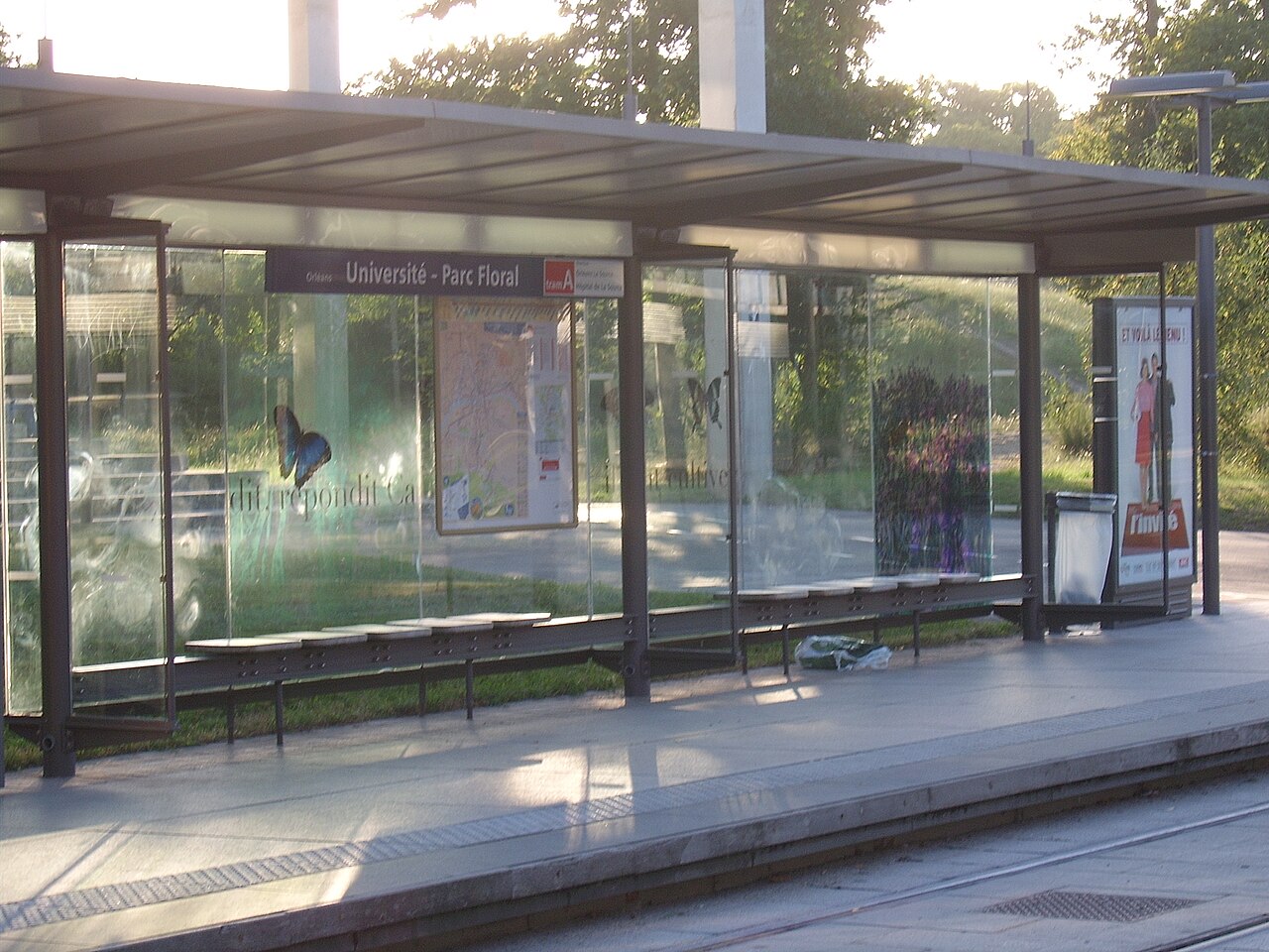 Arrêt de tram ou de bus moderne avec abri vitré, affiches publicitaires et panneaux d’information, près d’un trottoir en plein jour. Le décor montre une zone urbaine calme. Coût de la vie à orléans est évoqué par le contexte.