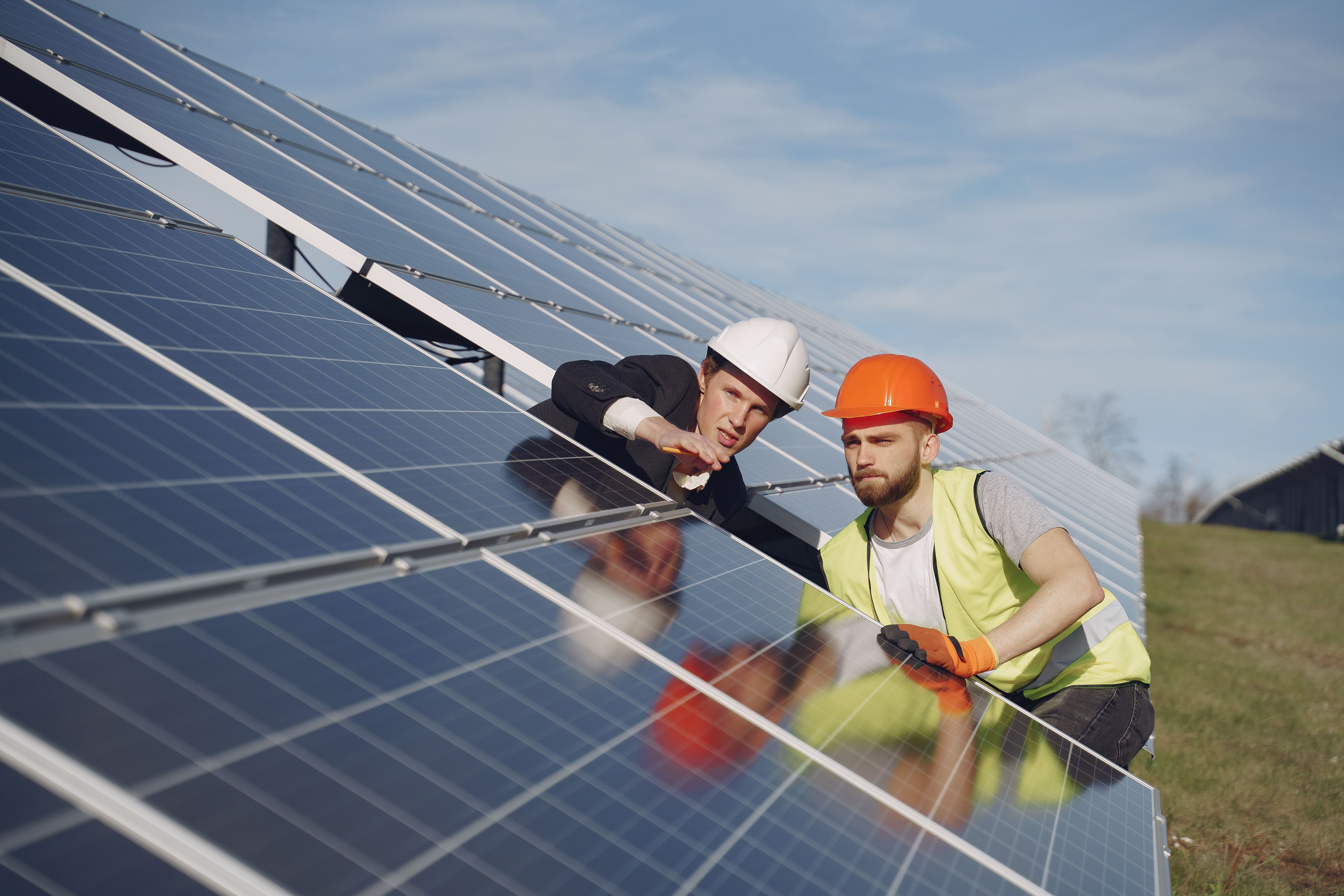 Two workers in hard hats install or inspect solar panels on a sloped array in a field, part of a commercial solar installation process.