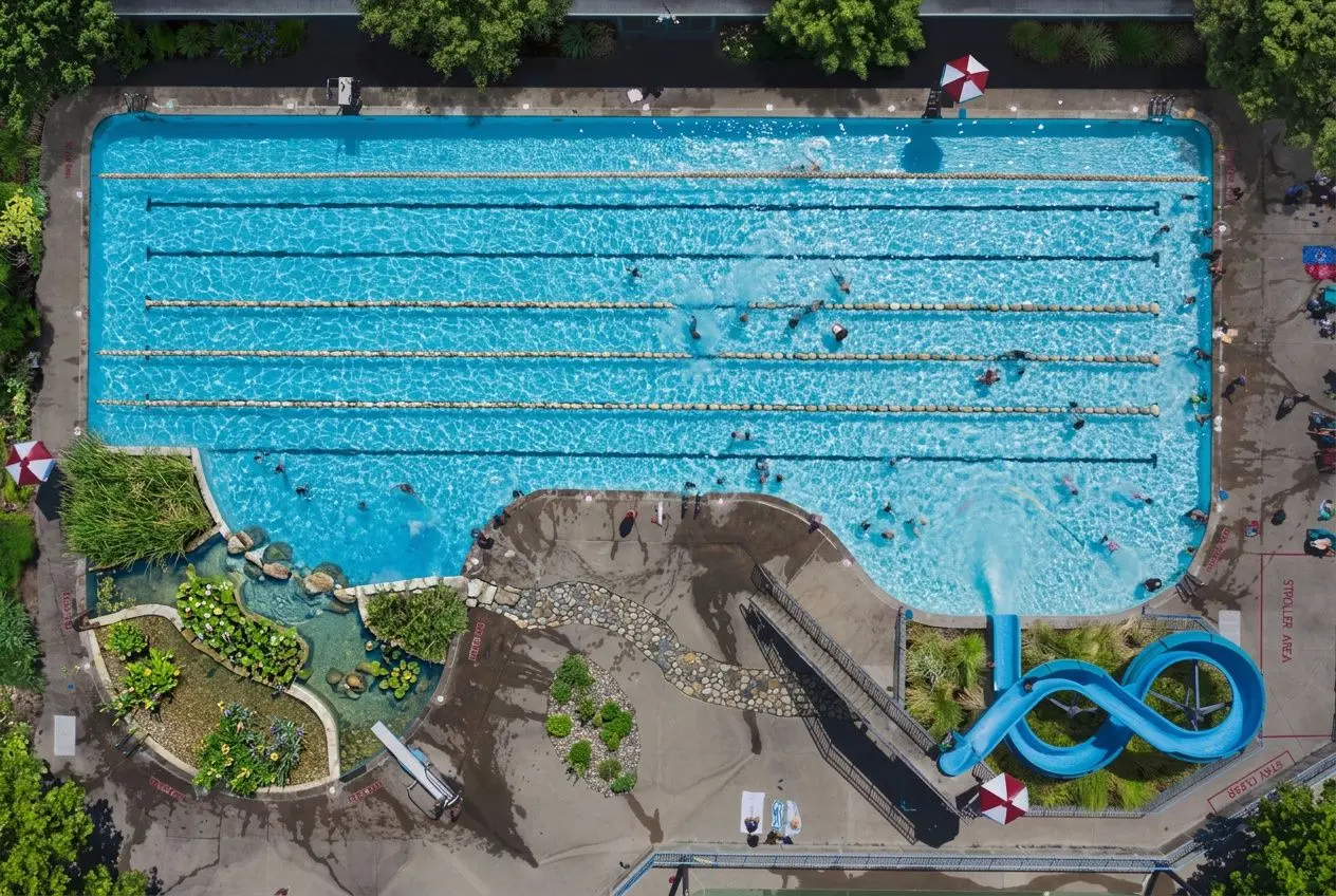 Vista aérea de una piscina pública con varias líneas de nado y tobogán azul en el extremo; áreas de césped y jardines alrededor. Describe la filtración piscina natural? Integrate keyword naturally: "filtración piscina natural" fits? The image shows una piscina con sistemas de filtración natural no químico. Let's craft alt: "Piscina pública con canaletas de nado y tobogán azul; zona de jardinería y filtración piscina natural visible en su entorno." Ensure 50-125 chars. Let's count: "Piscina pública con canaletas de nado y tobogán azul; zona de jardinería y filtración piscina natural visible." That's long. Remove some: "Piscina pública con canaletas de nado y tobogán azul; jardines y filtración piscina natural visible." Good. Piscina pública con canaletas de nado y tobogán azul; jardines y filtración piscina natural visible.