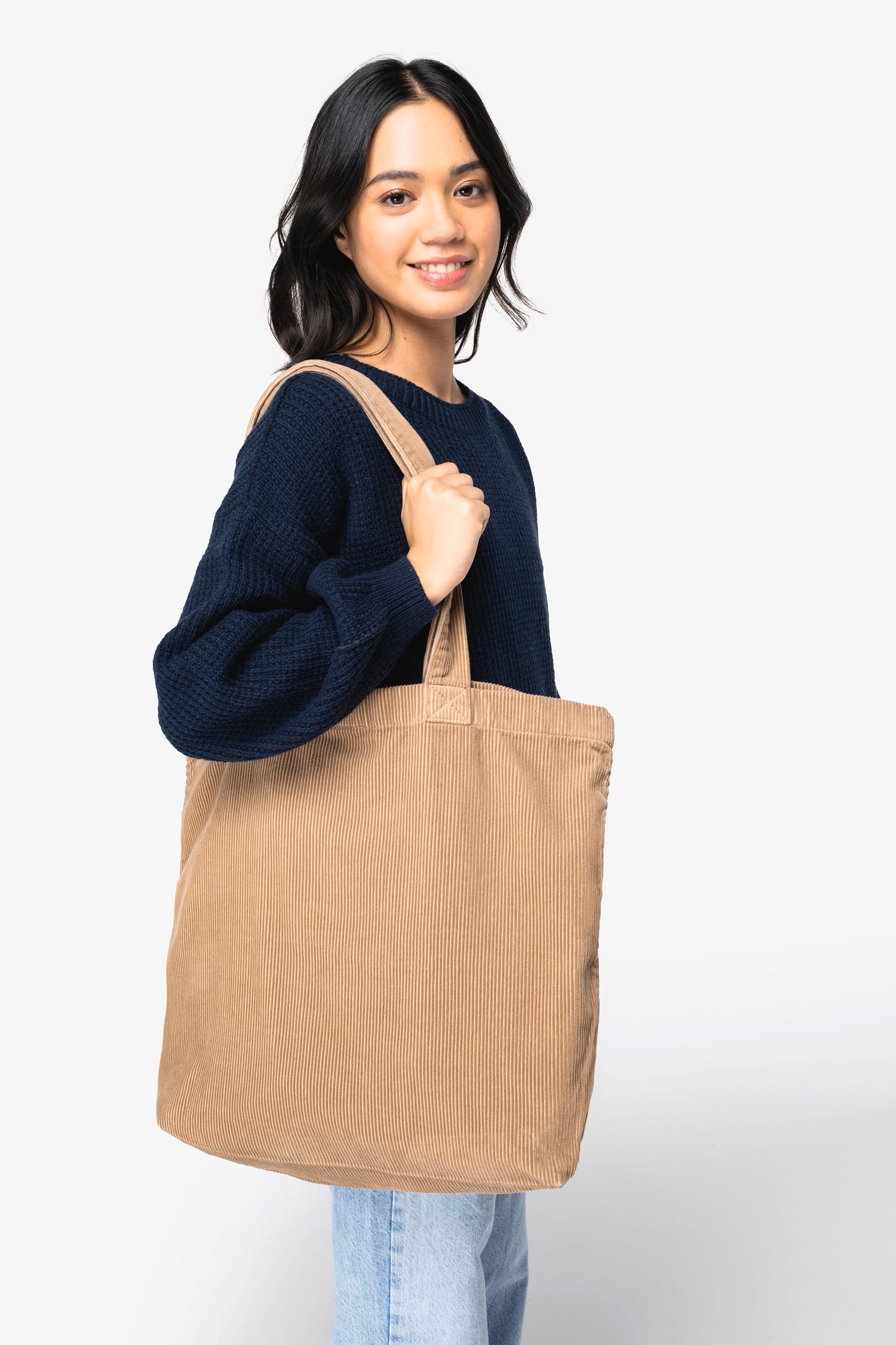 Femme portant un tote bag brodé personnalisé en tissu beige, veste bleu marine et jeans, sur fond blanc.