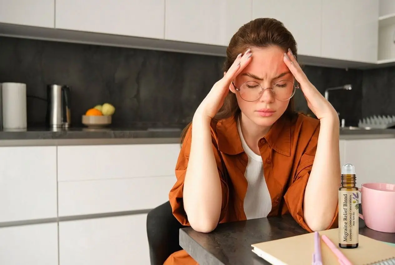 une femme assise à une table, se tenant la tête avec les doigts sur les tempes, manifestant un mal de tête, avec un flacon d’huile essentielle sur le bureau et des objets de cuisine en arrière-plan