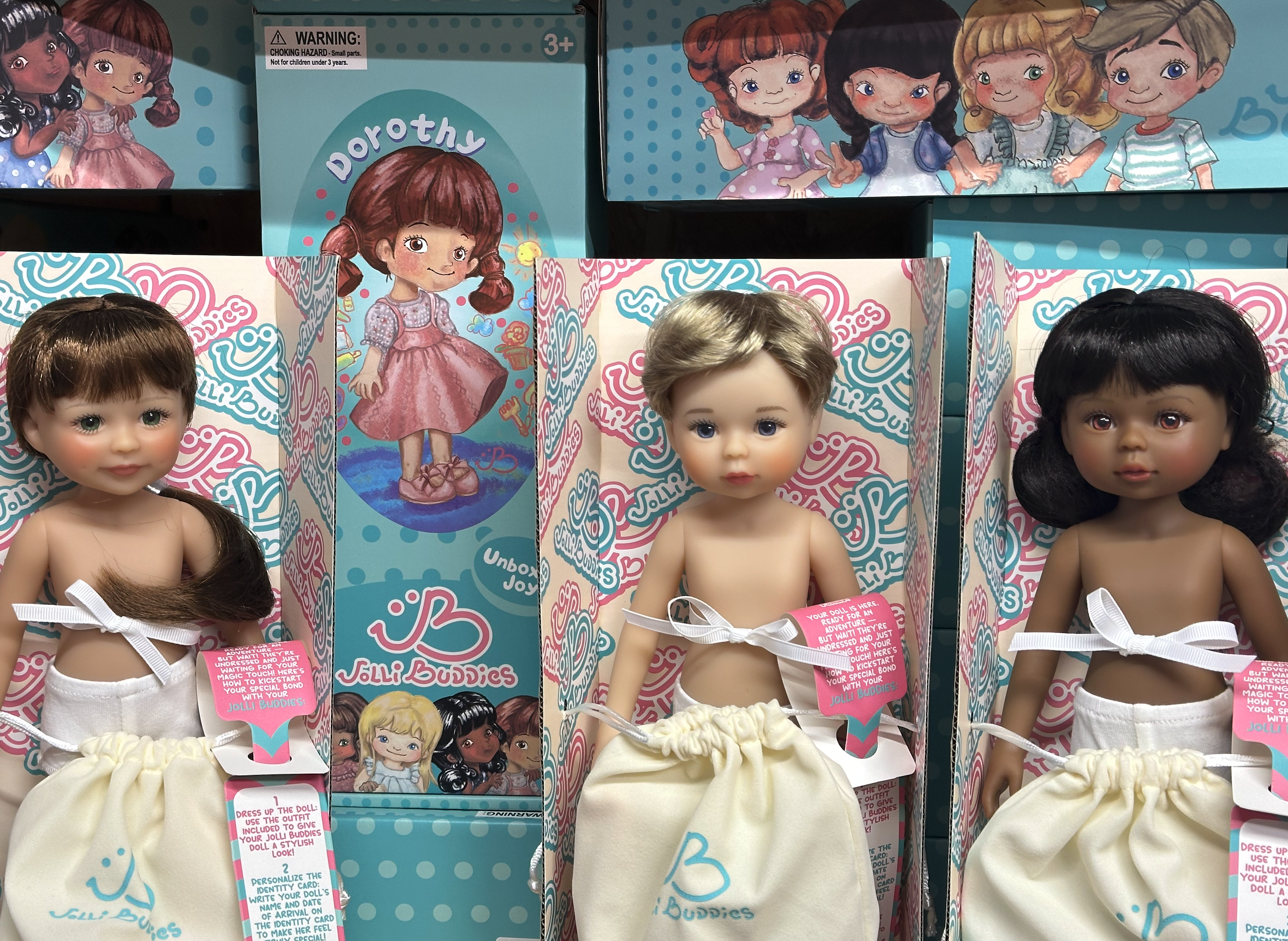 Poupées Jolli Buddies en boîtes colorées, boxes turquoise et roses affichant des visages d’enfants; tenues blanches avec rubans. Poupées Jolli Buddies en boîtes colorées, boxes turquoise et roses affichant des visages d’enfants; tenues blanches avec rubans.