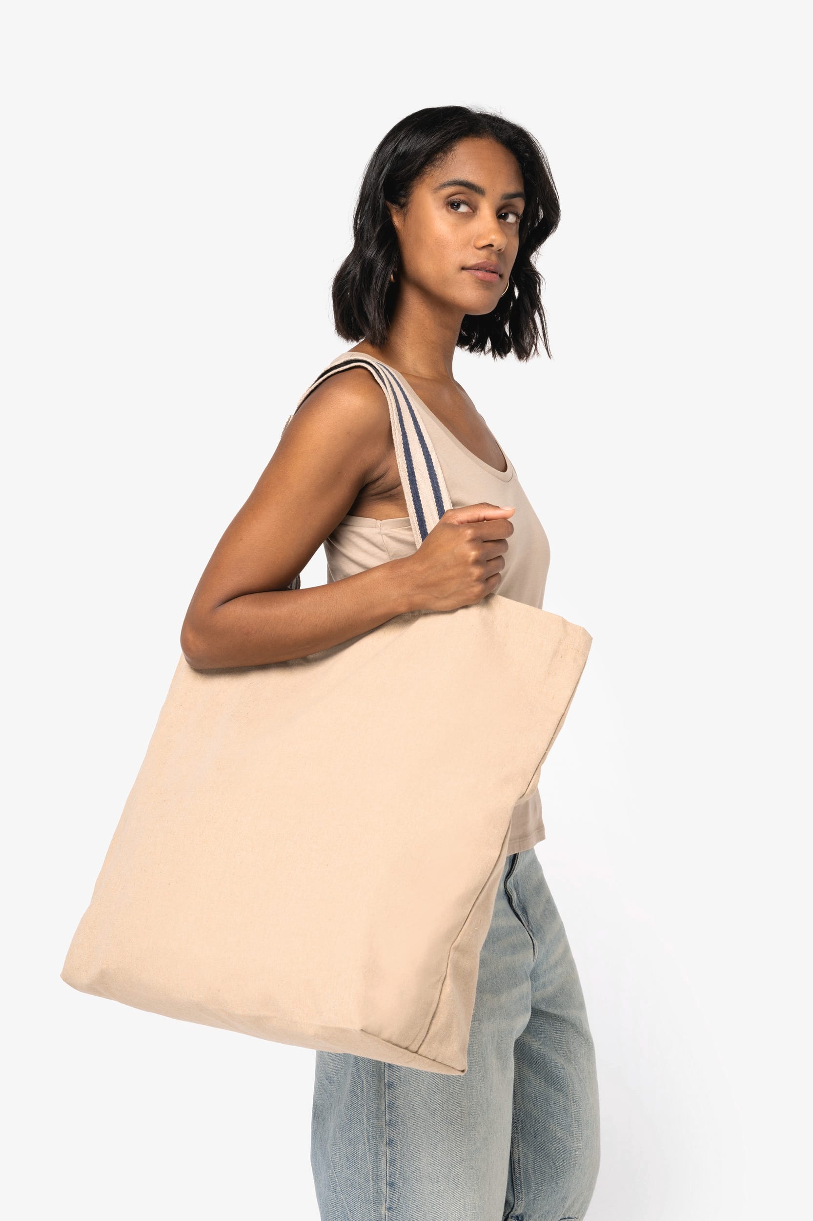 Tote bag beige simple portée sur l'épaule par une femme, porté par une sangle rayée, sans motif visible. Tote bag brodé personnalisé pour pros et associations.