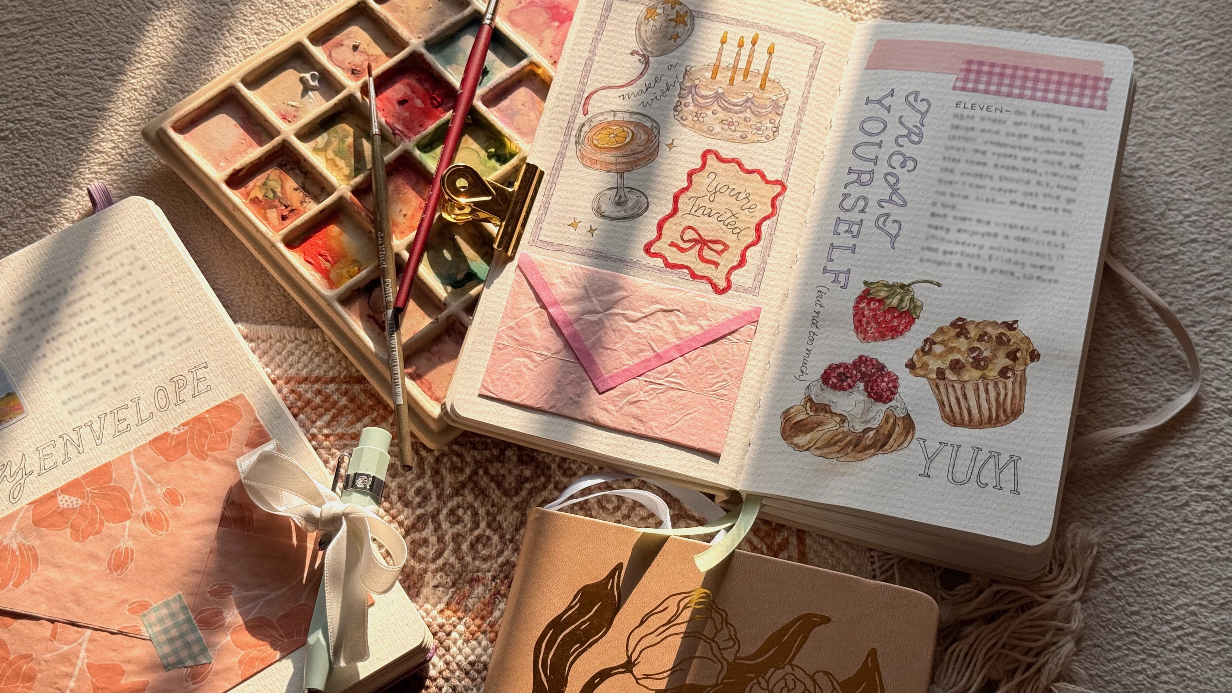 A flat lay of watercolor art supplies and journals: a paint palette, brushes, clips, and an illustrated open notebook with cake and fruit drawings, pastel envelopes, and a peach-toned journal. A flat lay of watercolor art supplies and journals: a paint palette, brushes, clips, and an illustrated open notebook with cake and fruit drawings, pastel envelopes, and a peach-toned journal.