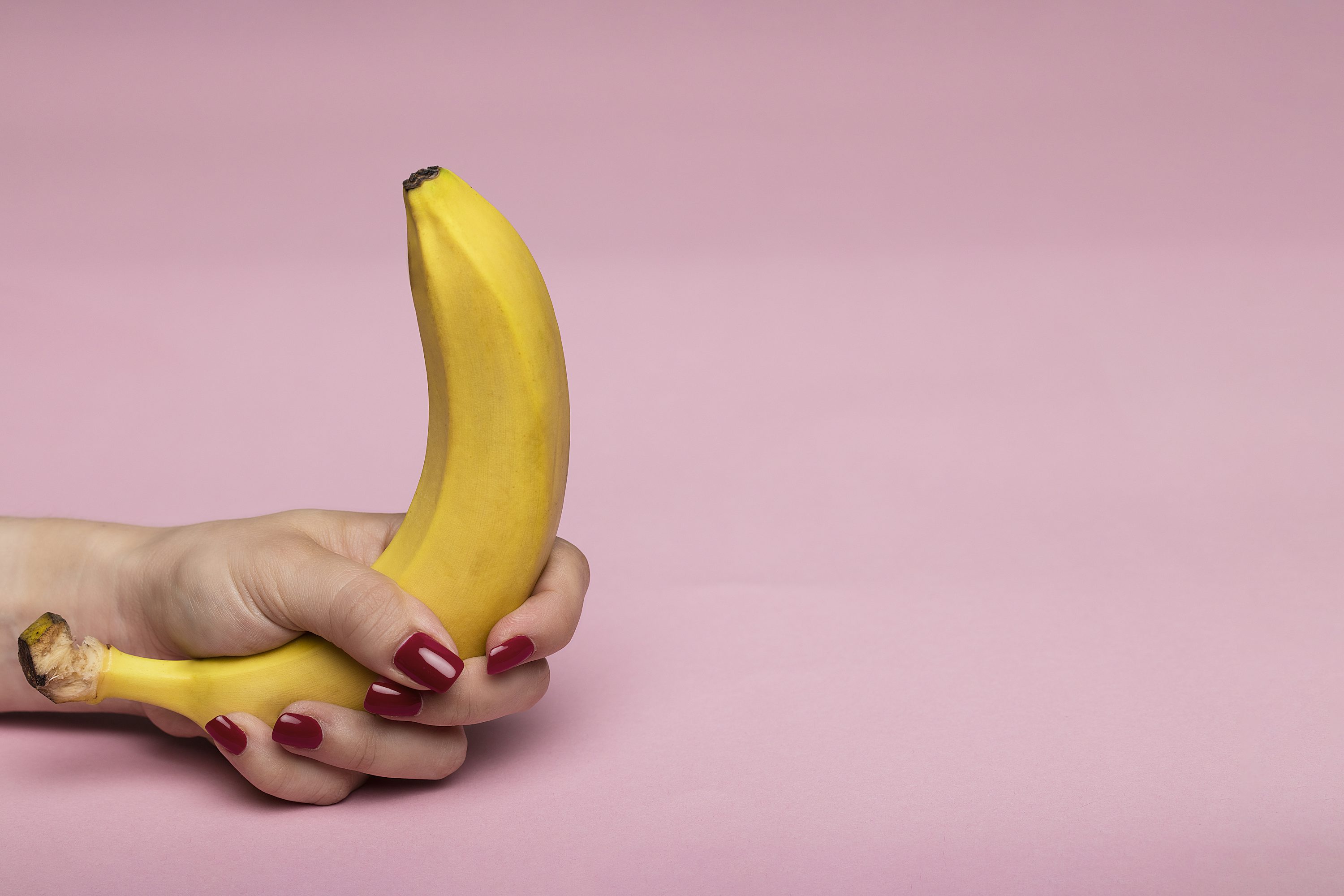 Main extrémité d'une main tenant une banane sur fond rose, avec ongles rouges et pose suggestive.