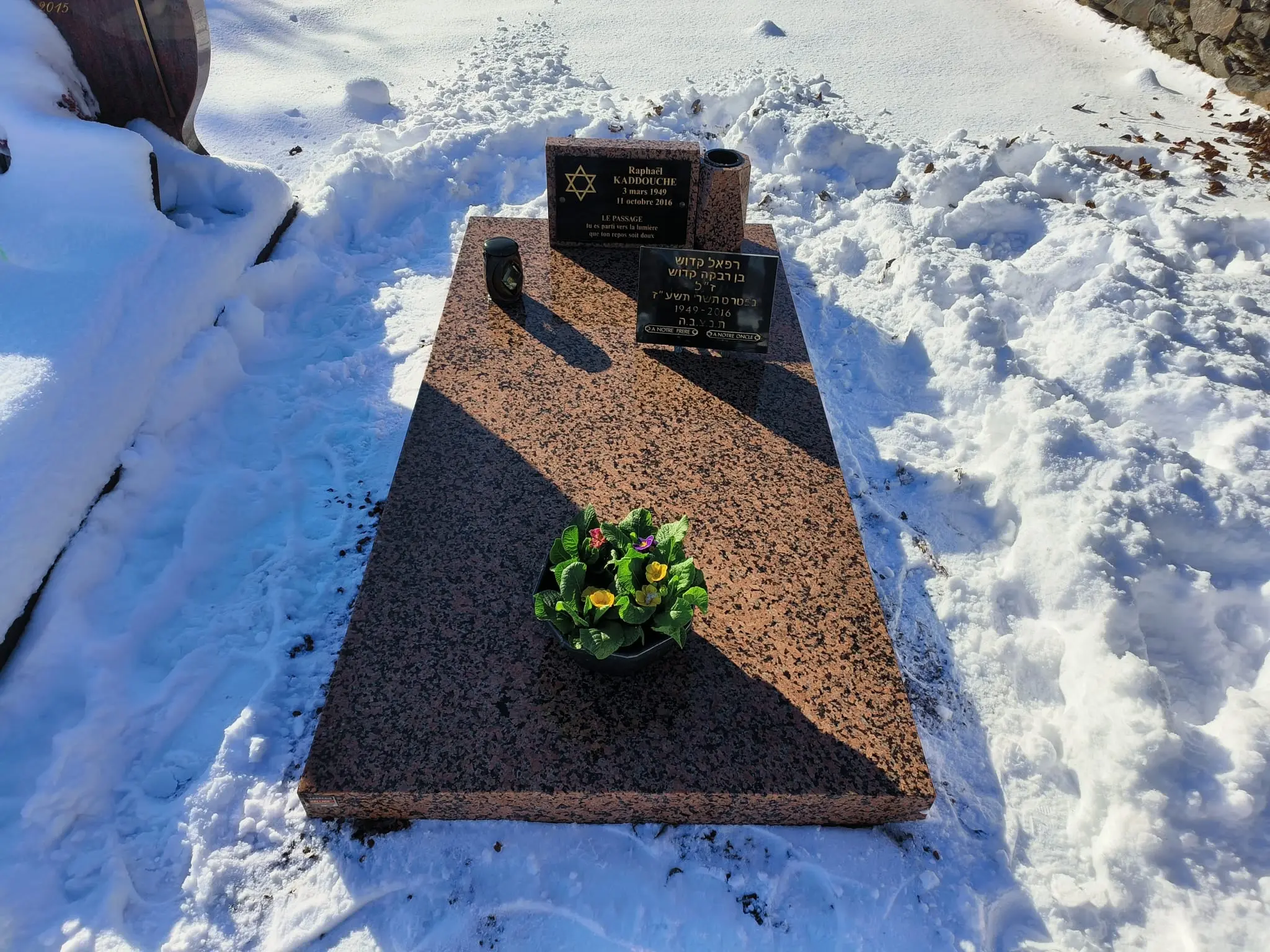 Pierre tombale en granit rouge avec petites plaques noires, fleuri devant et neige autour, en inscription multilingue et symbole étoile.