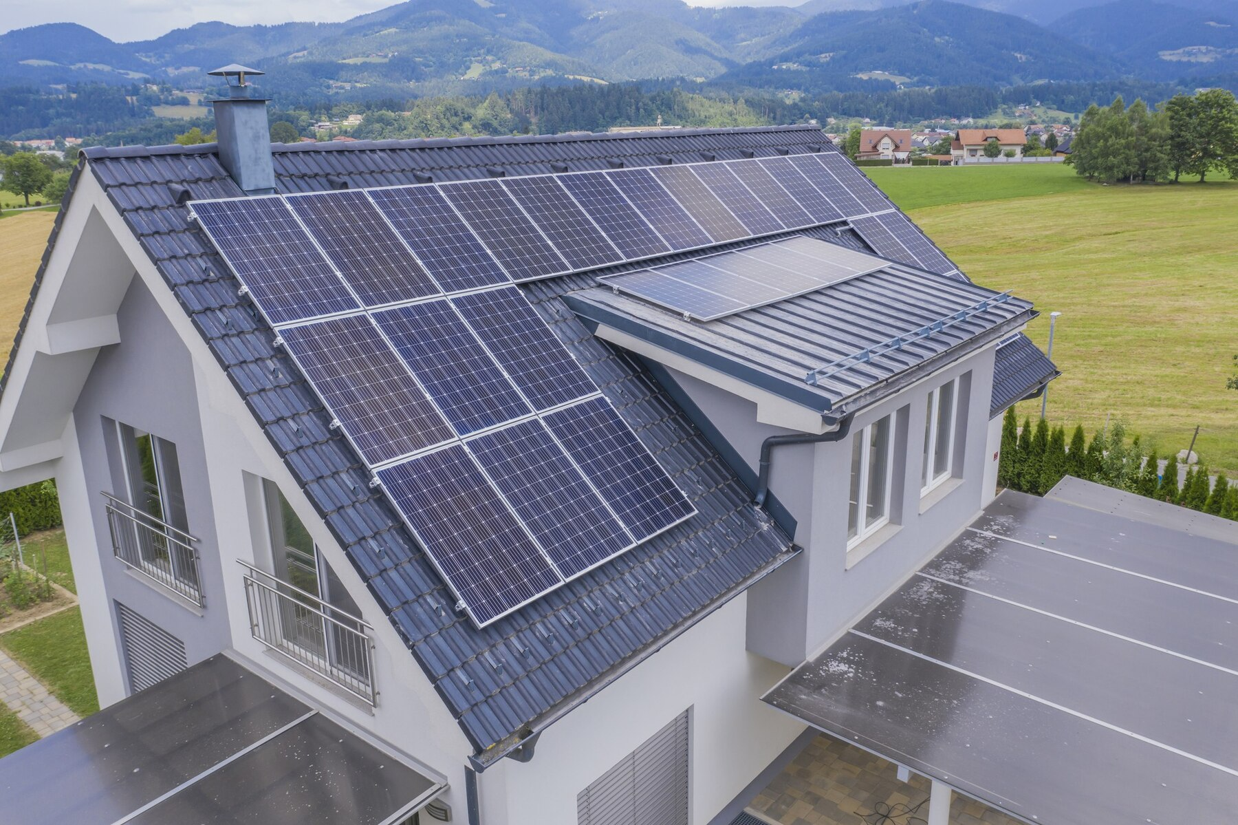 Rooftop solar panels cover a sloped roof beside a modern two-story house with a scenic mountainside backdrop.