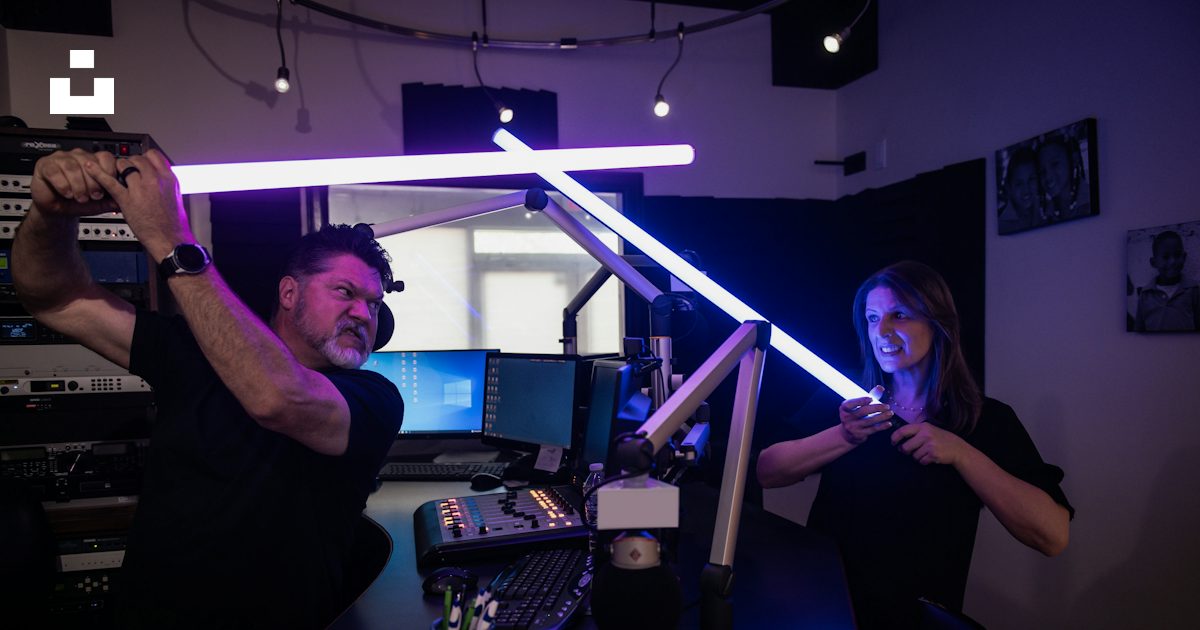 Deux personnes dans un studio, l’une manie un éclairage lumineux au-dessus d’un poste de travail audio et l’autre tient un