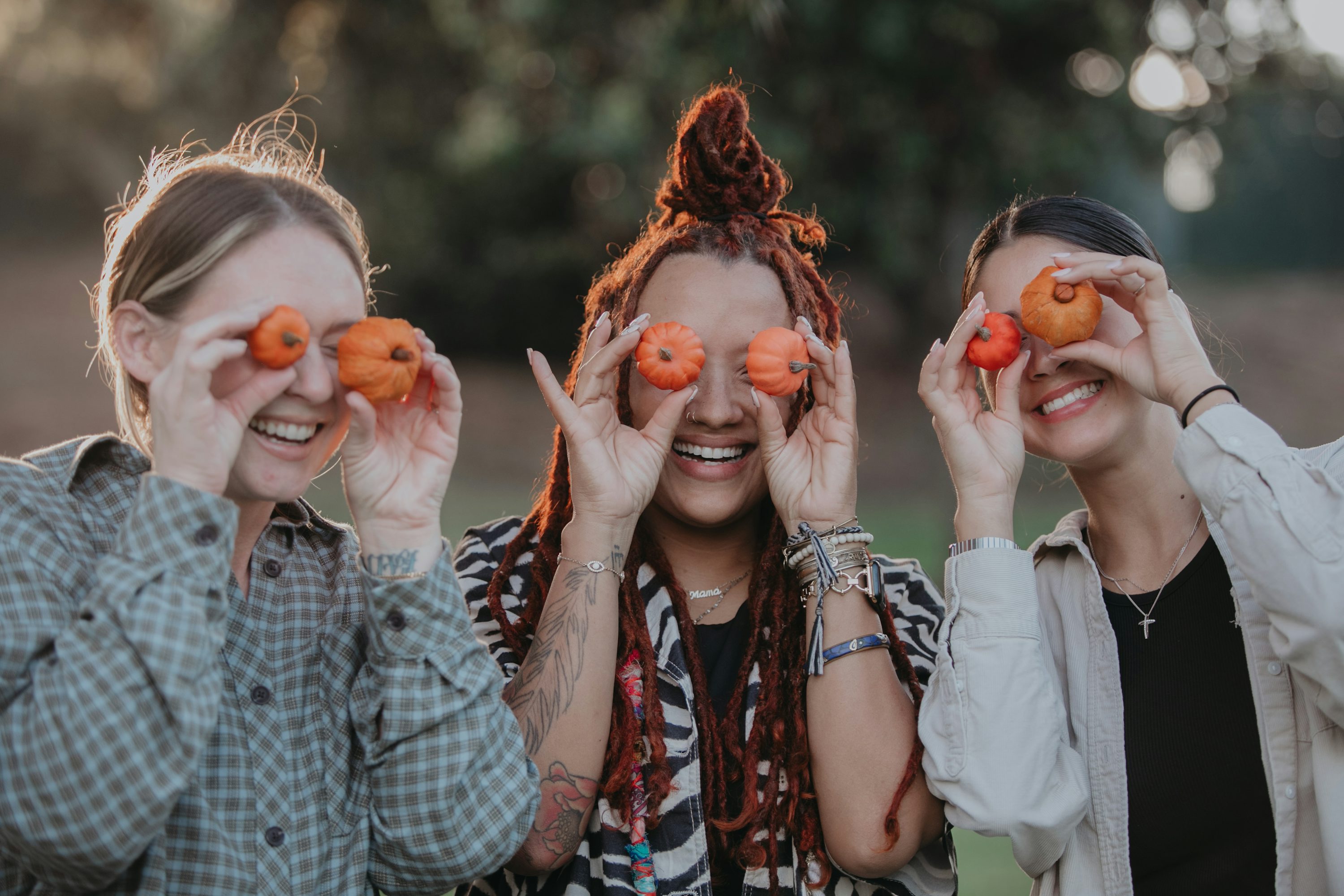Trois femmes souriantes jouent à cacher leurs yeux avec des miniatures orange vif, en plein air.